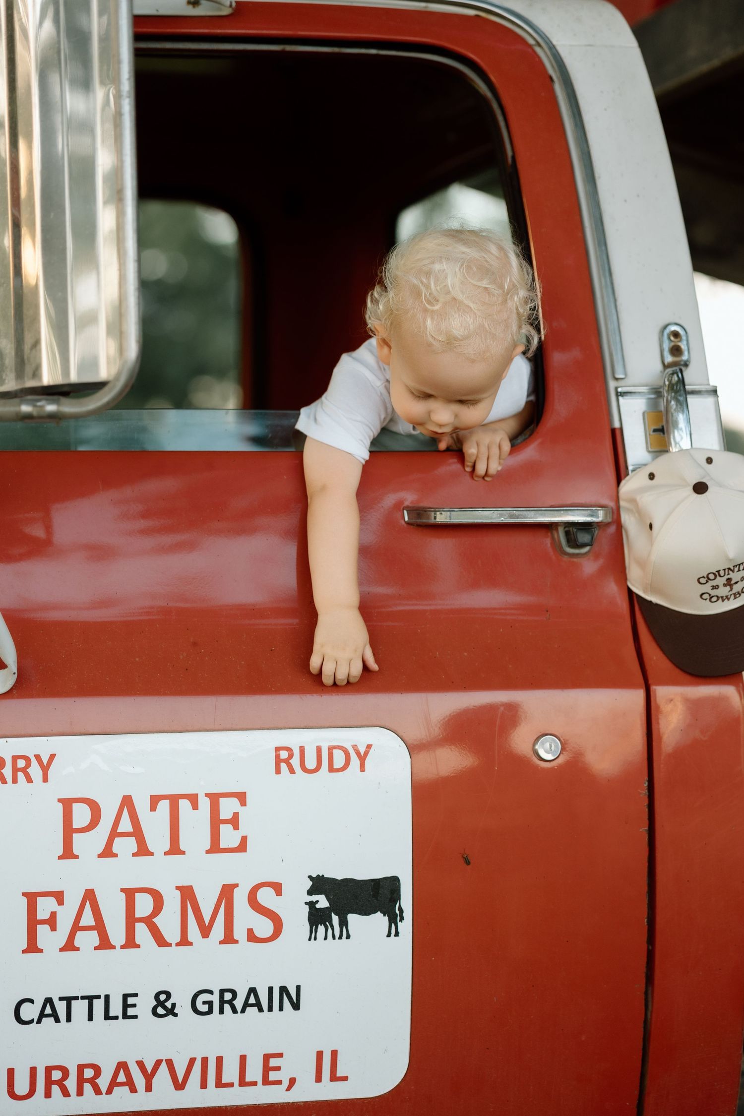 Curious toddler leans out window of vintage red Pate Farms truck door.