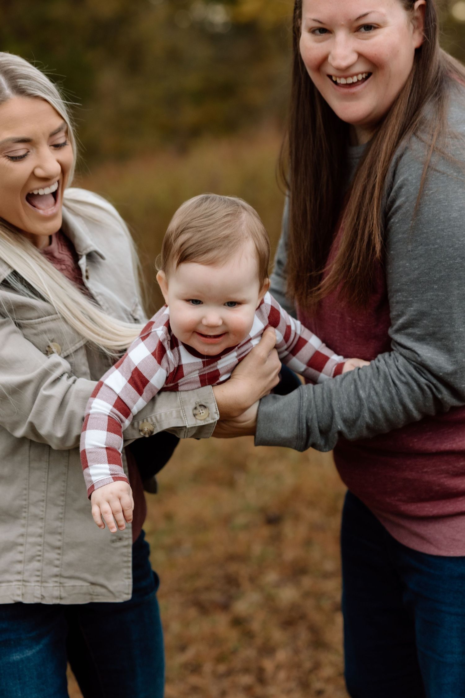Two women laughing joyfully while holding a smiling baby in a plaid shirt outdoors in autumn.