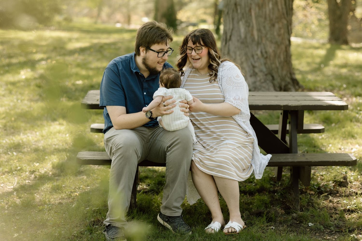 Young couple sits at picnic table in park, lovingly gazing at their newborn baby in soft natural light.