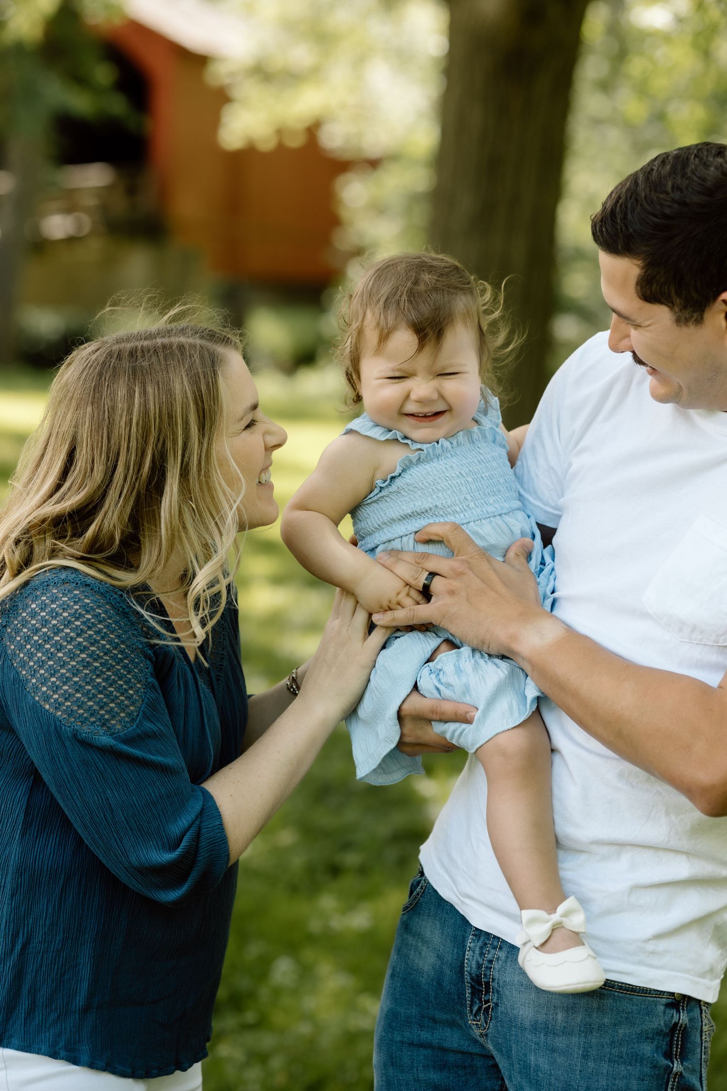 Smiling parents hold their laughing baby girl in a blue dress during an outdoor family photo session in a park.