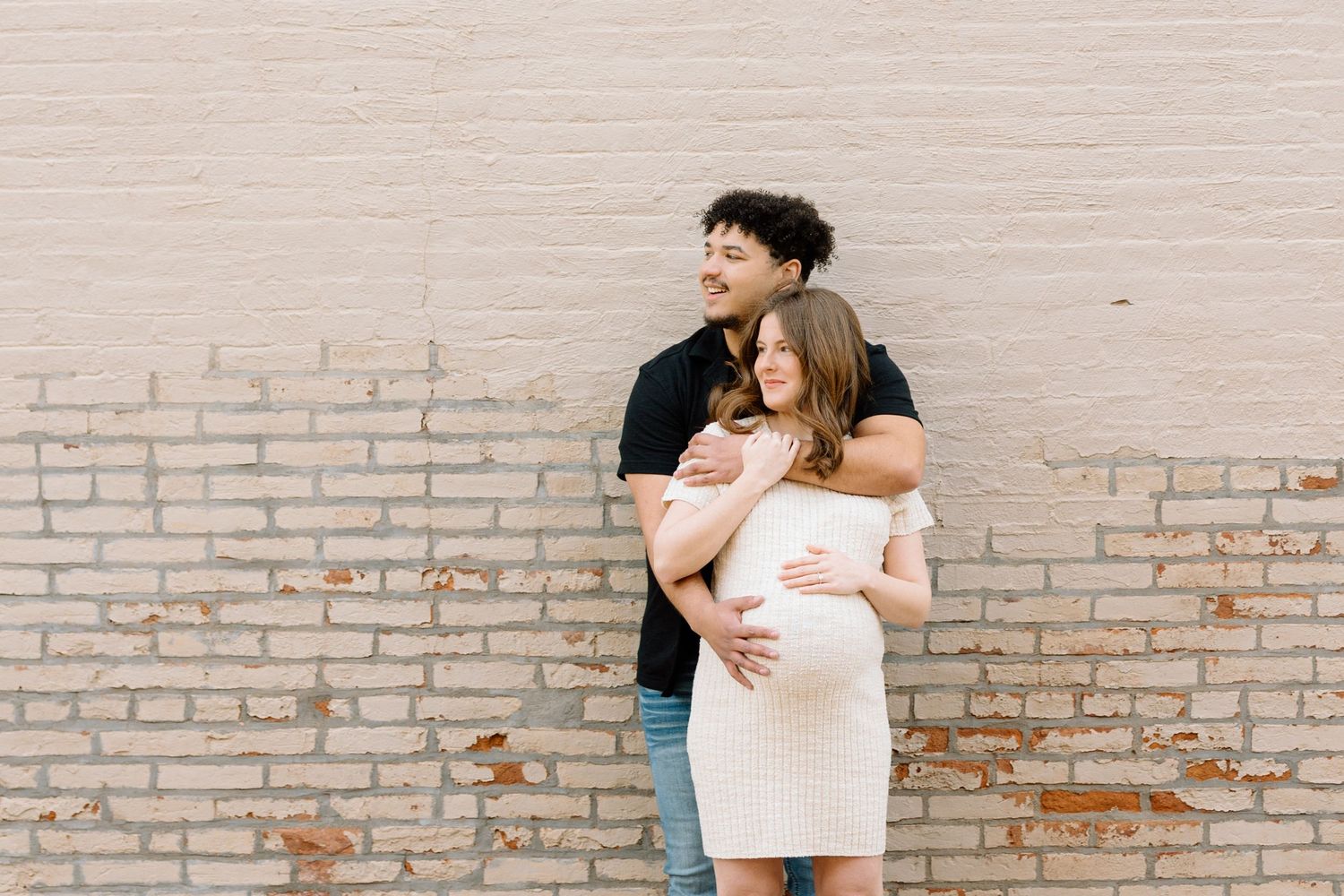 Couple embracing during maternity photoshoot against light brick wall, man hugging pregnant woman in cream dress.