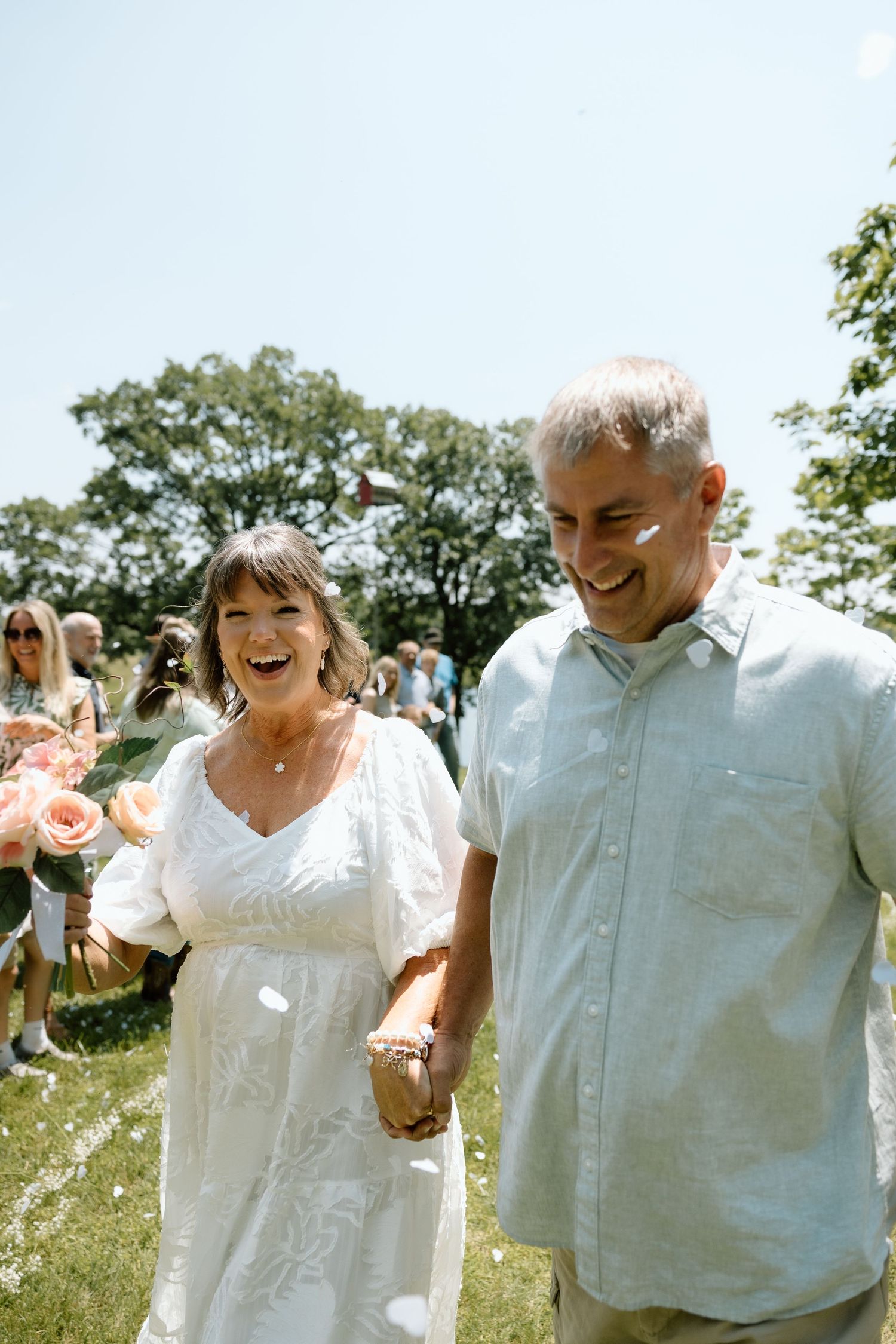 A joyful older couple walks hand in hand through a sunny outdoor wedding ceremony surrounded by guests.