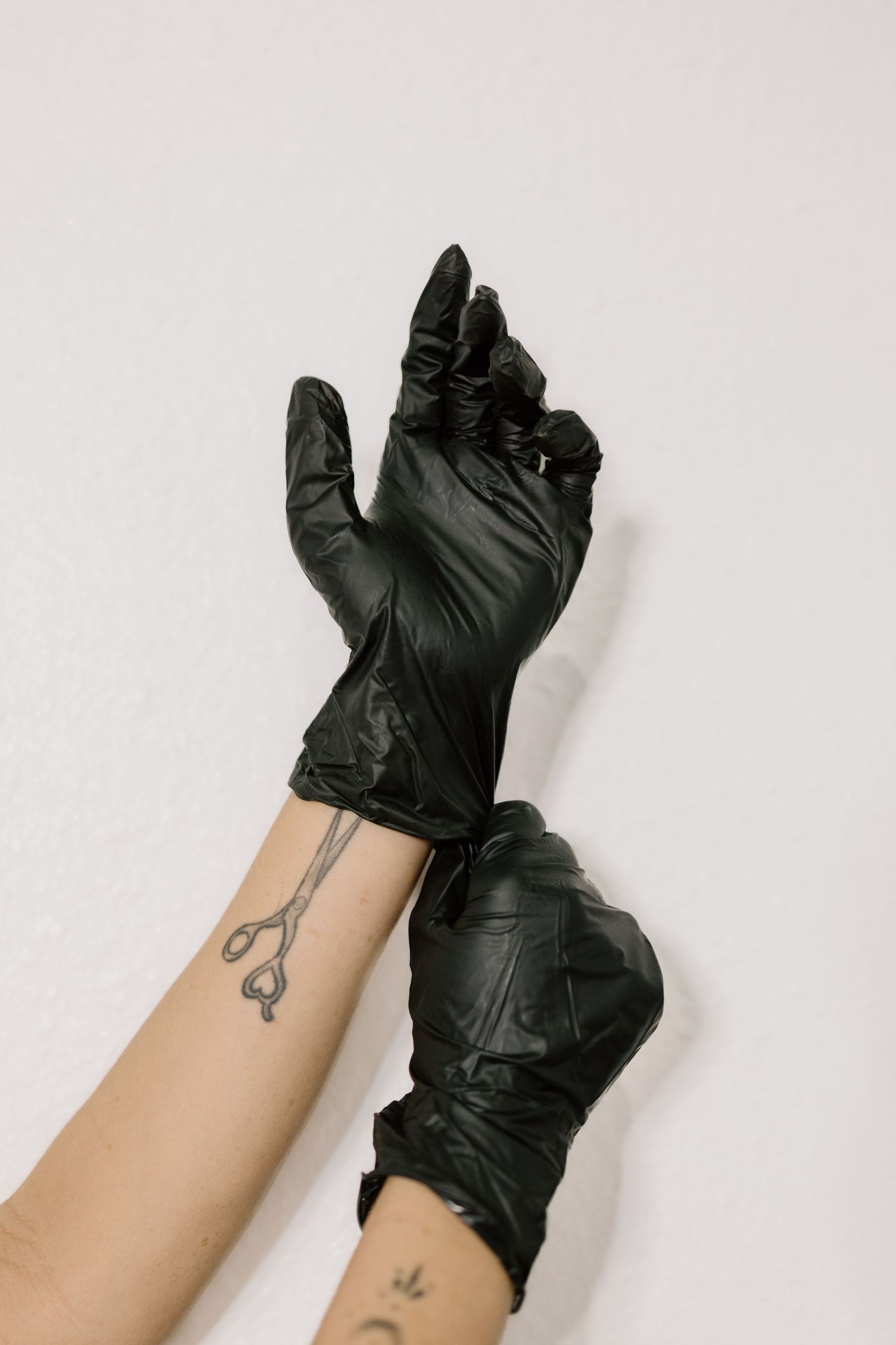 Hands wearing black latex gloves with scissors tattoo on forearm against white background.
