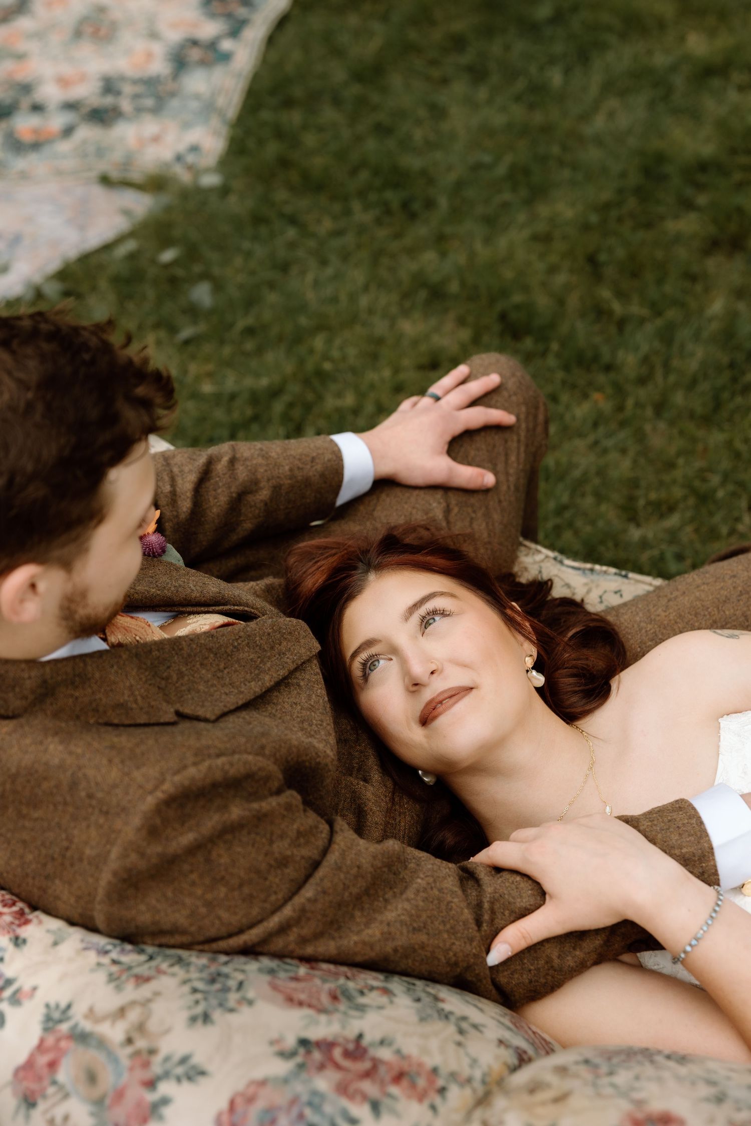Bride in white strapless dress gazes up at groom in brown suit while lying on floral cushion.