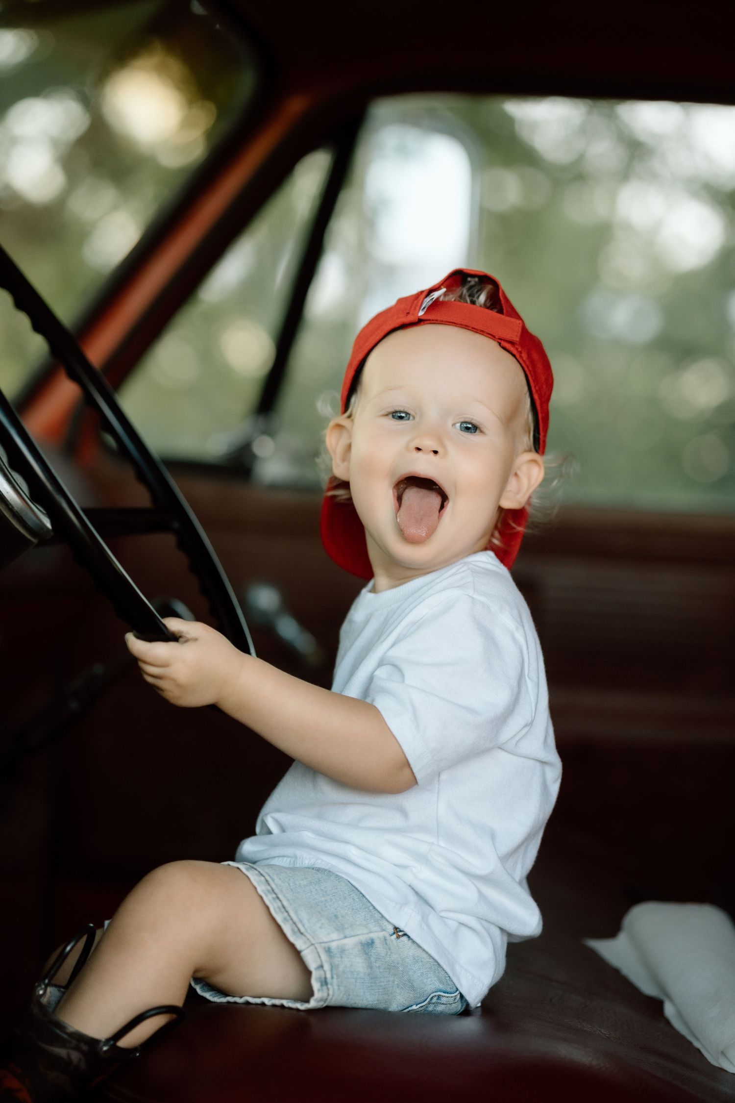 Laughing toddler in red cap and white tee holds steering wheel of vintage red truck.