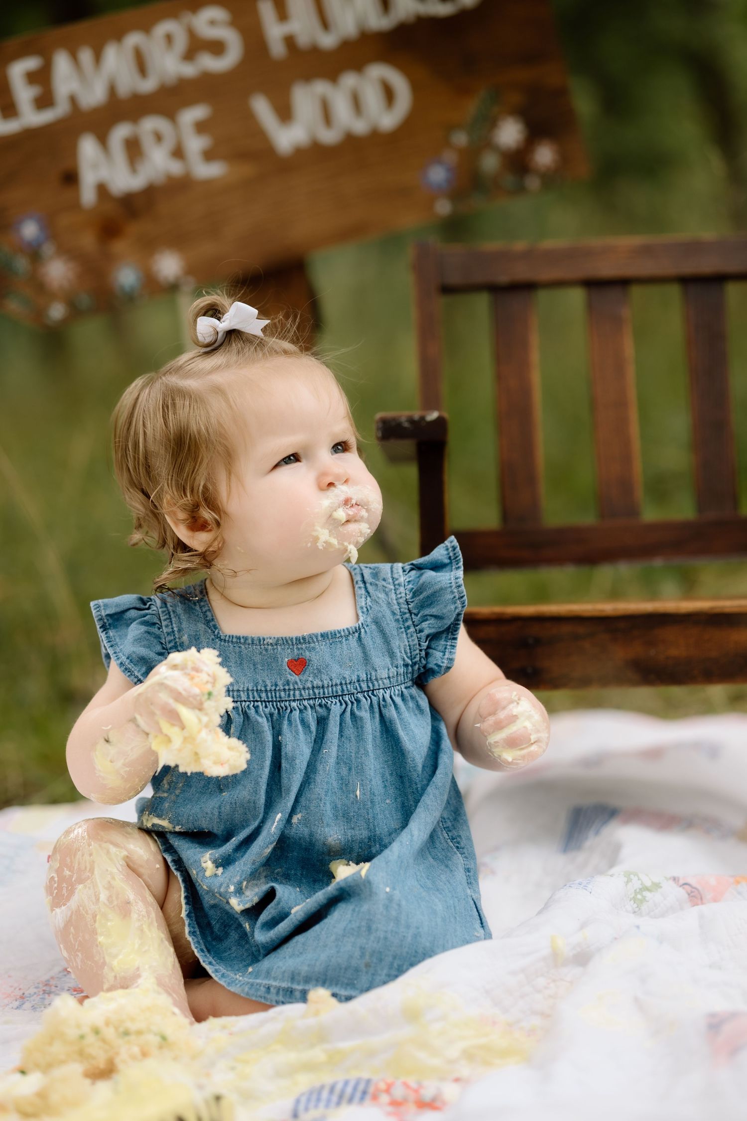Cute baby girl in denim dress eating birthday cake with frosting on her face outdoors.