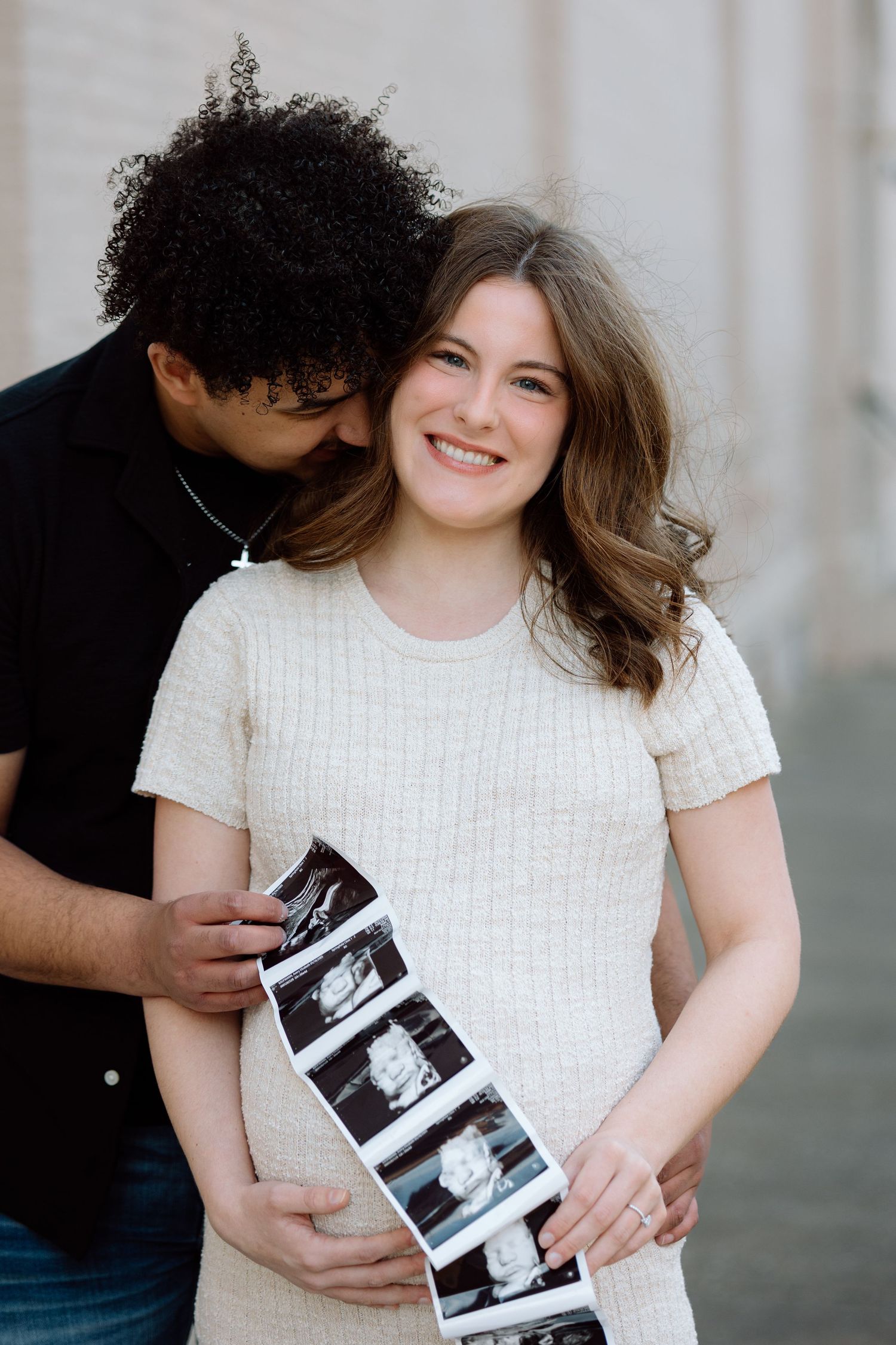 Happy pregnant couple displaying ultrasound photos while man kisses womans cheek during maternity session.