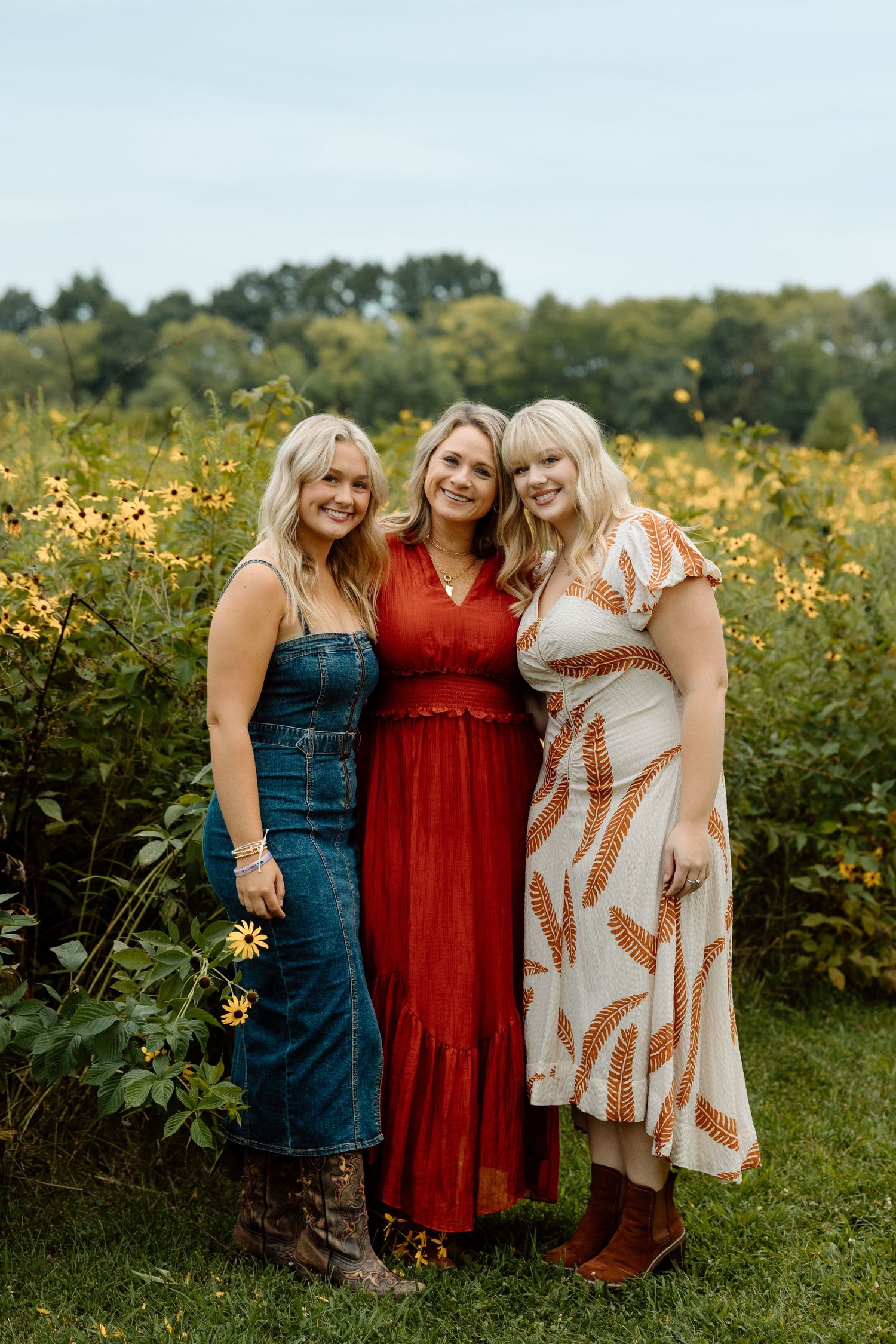 Three young women in colorful dresses smiling together in front of a bright wildflower field.