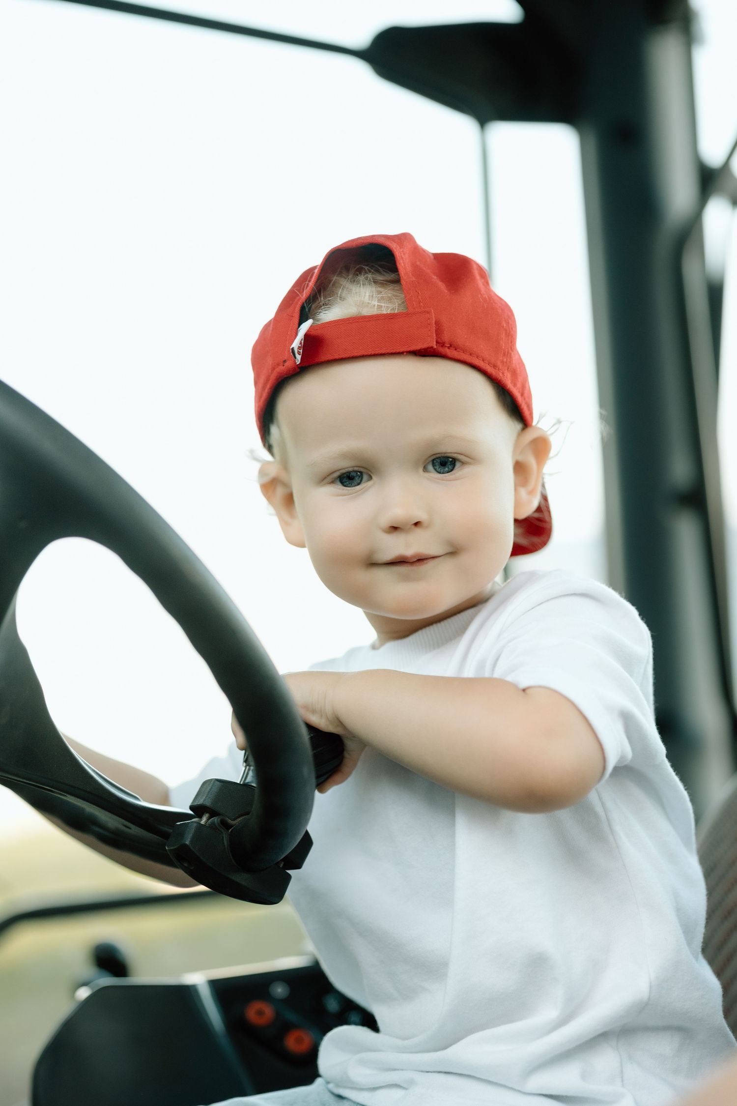 Smiling toddler boy in red cap sitting at a tractor steering wheel on a farm.