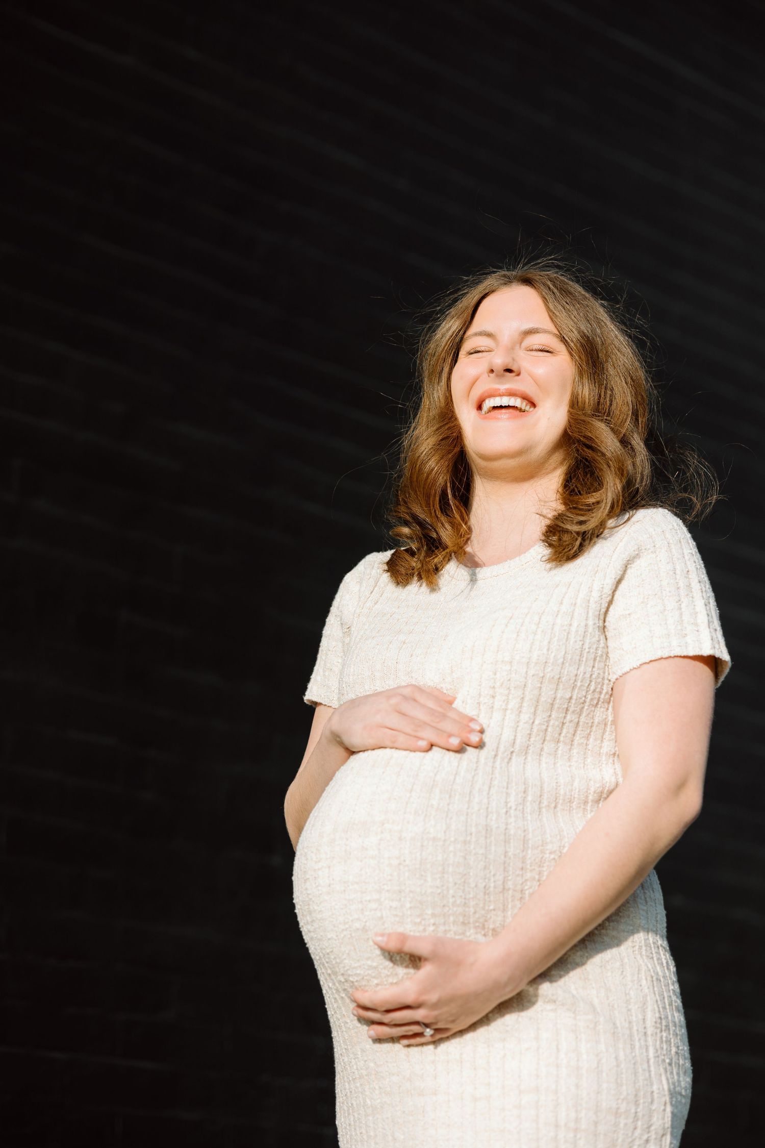 Joyful pregnant woman in white dress laughing while cradling her baby bump against a dark background.