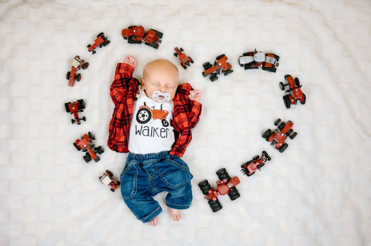 Baby named Walker in flannel shirt and jeans lies on white fur surrounded by orange toy tractors arranged in a heart shape.