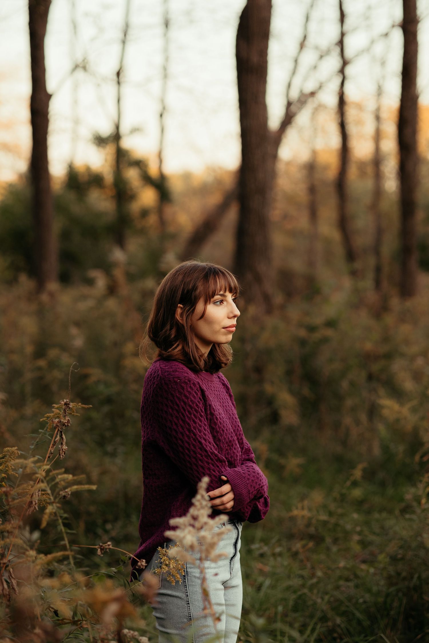 Young woman in purple knit sweater and jeans standing thoughtfully in autumn forest at golden hour.
