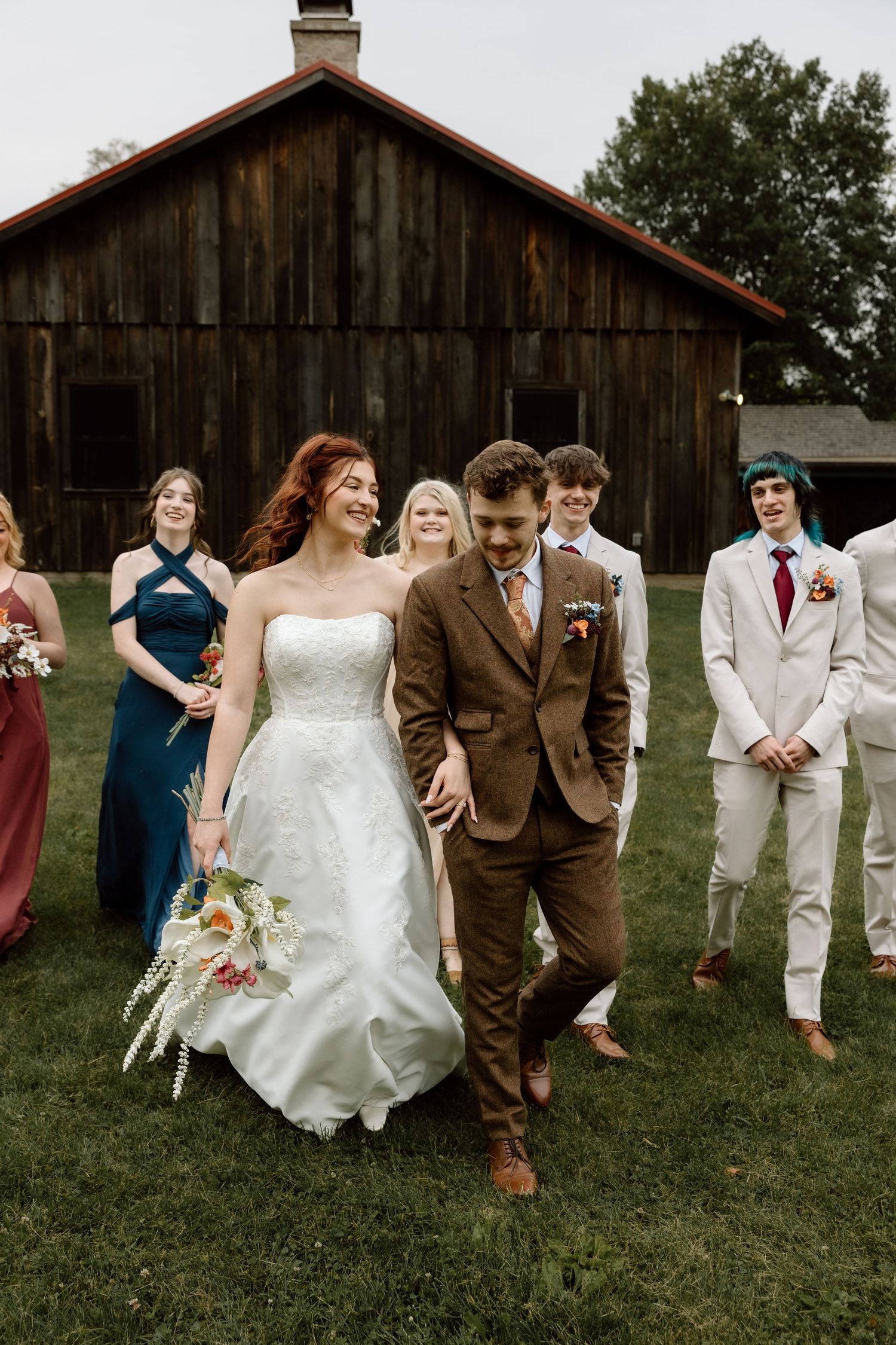 Bride and groom walk joyfully with wedding party in front of rustic barn venue.