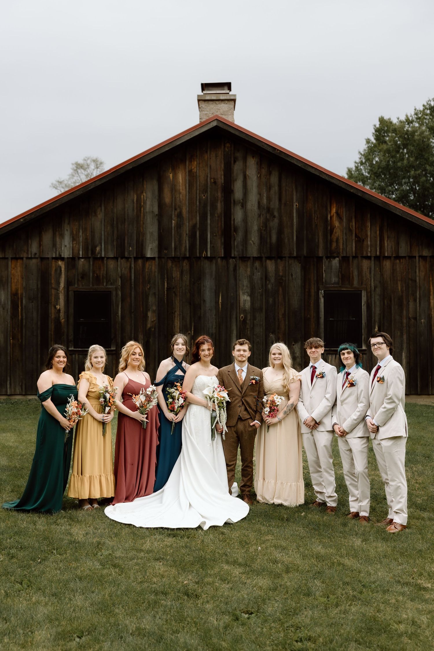 Wedding party poses before rustic wooden barn, bridesmaids in colorful gowns and groomsmen in neutral suits.