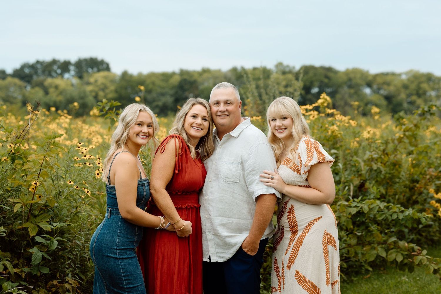 Smiling family of four posing closely together in a beautiful sunflower field on a cloudy day.