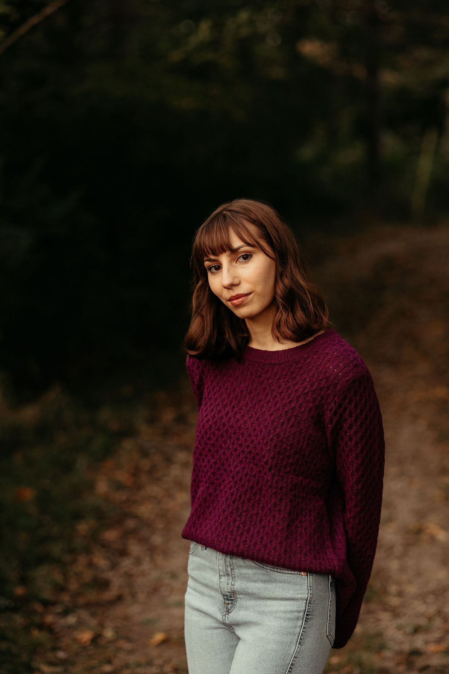 Young woman in purple knit sweater stands on a forest path looking directly at camera.