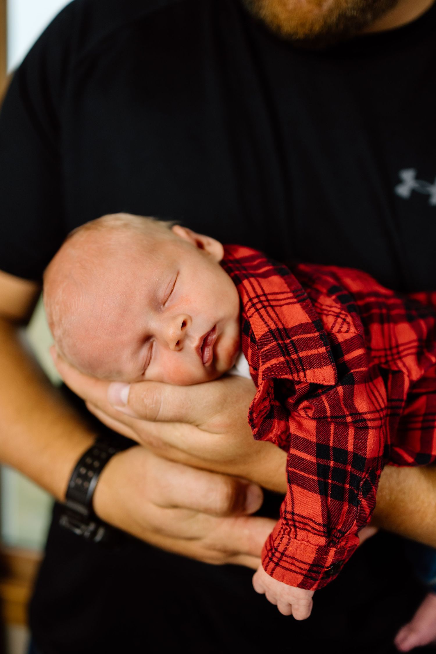 Close-up of a sleeping newborn baby in a red plaid shirt held gently in fathers hands.