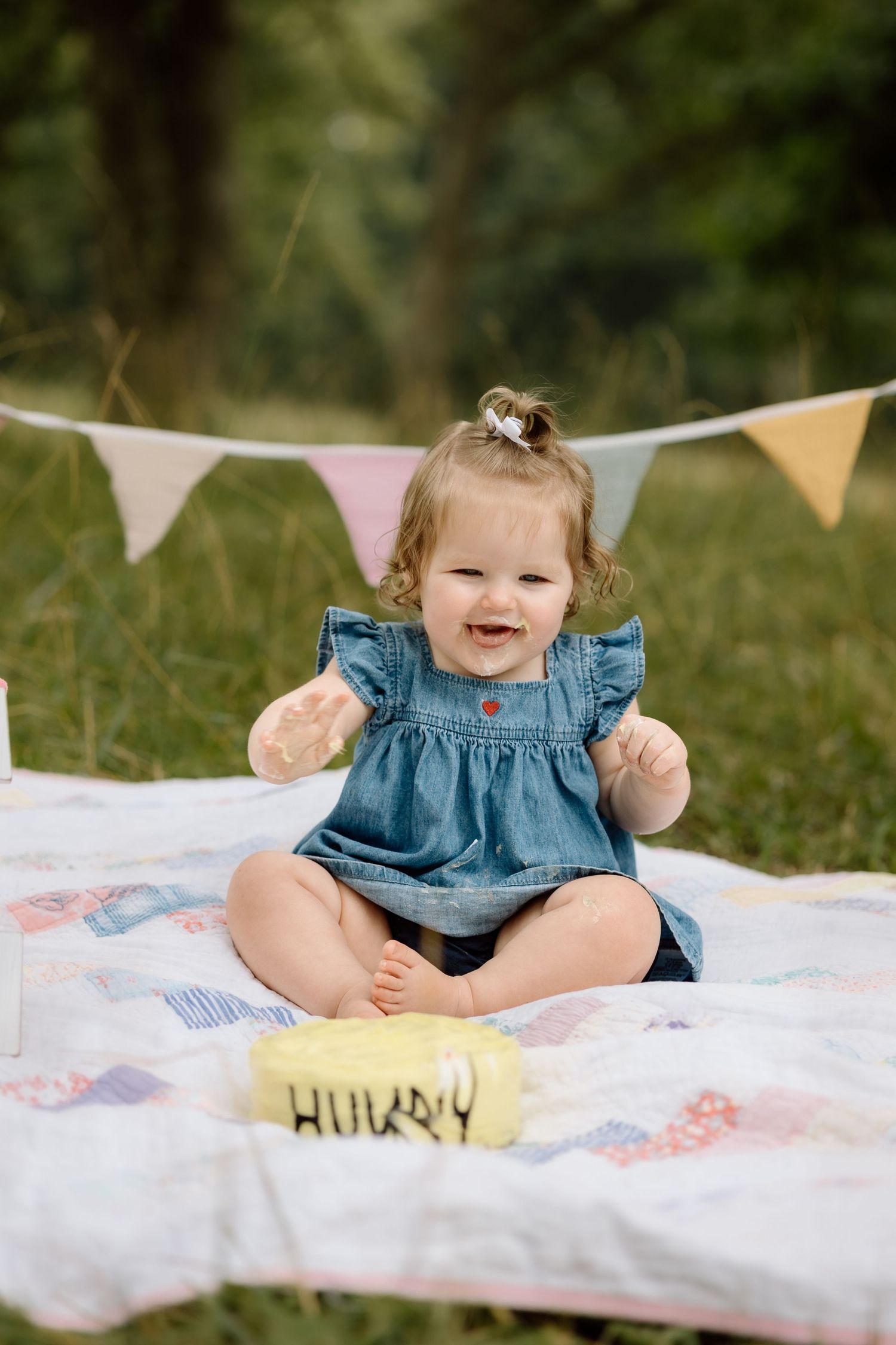 Joyful baby girl in denim dress clapping hands near yellow birthday smash cake on outdoor quilt.
