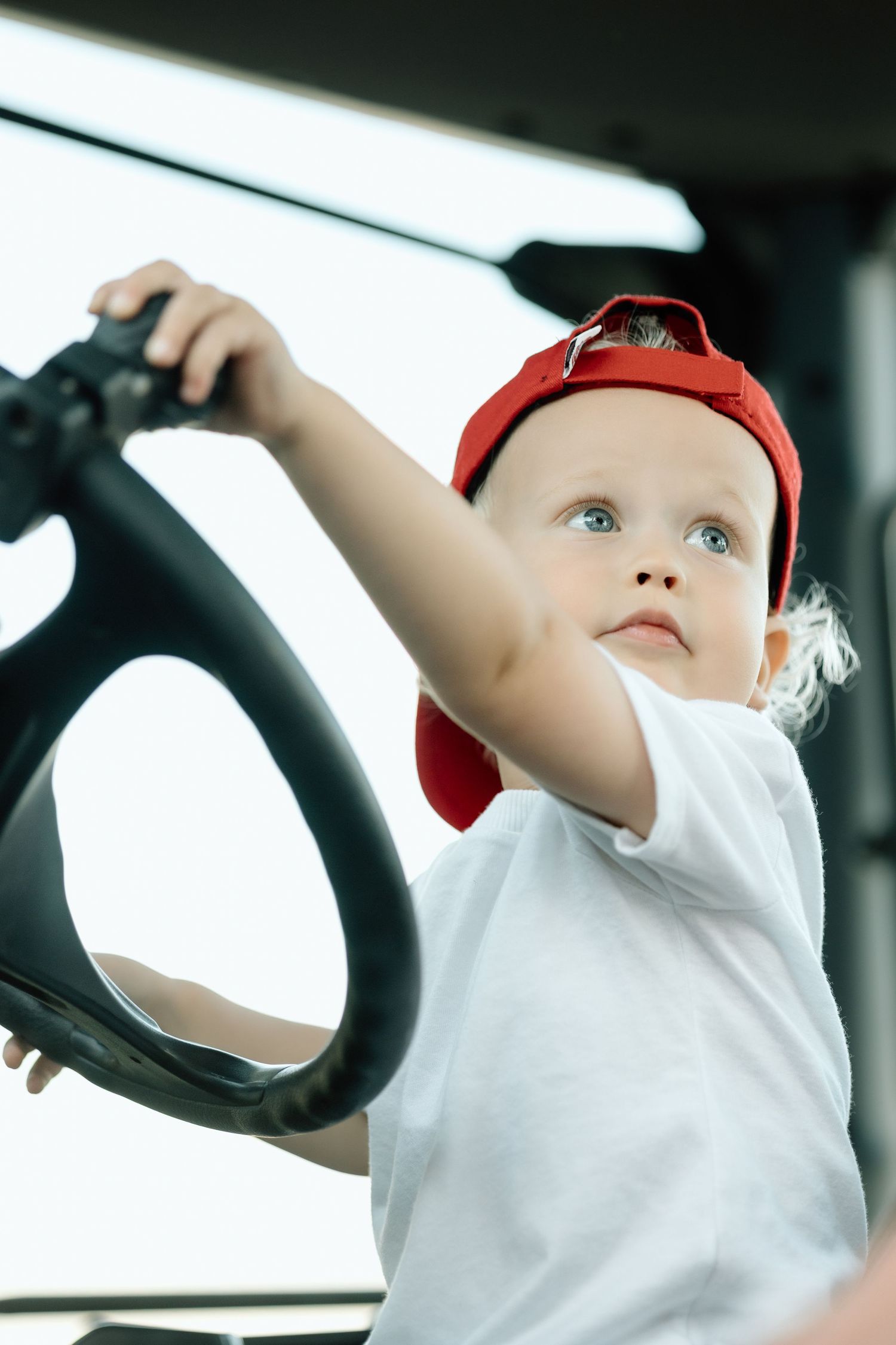 Curious toddler in red cap reaching for a tractor steering wheel inside a farm vehicle.