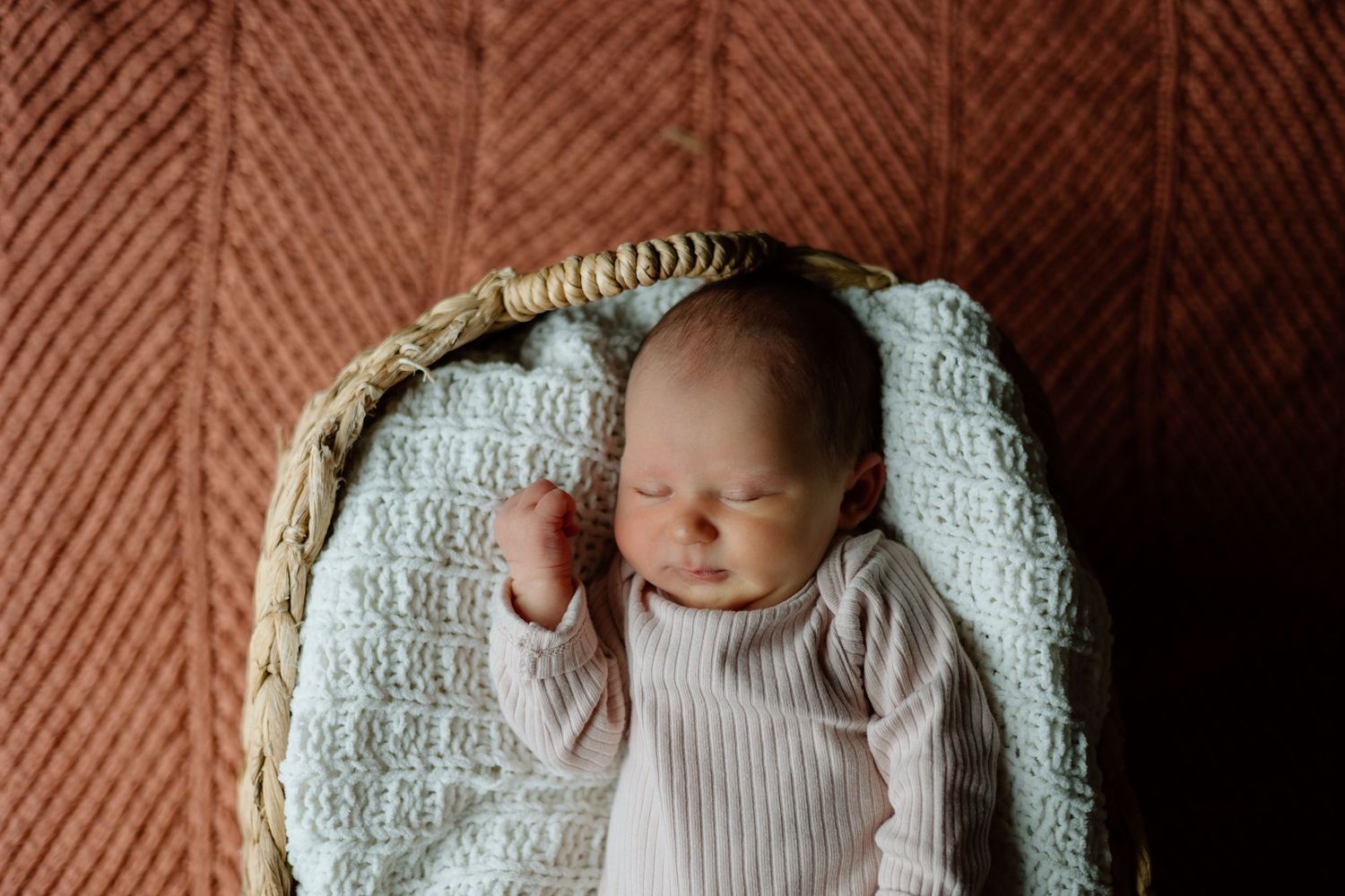 Sleeping newborn baby in pink onesie rests in wicker basket on rust knit blanket.