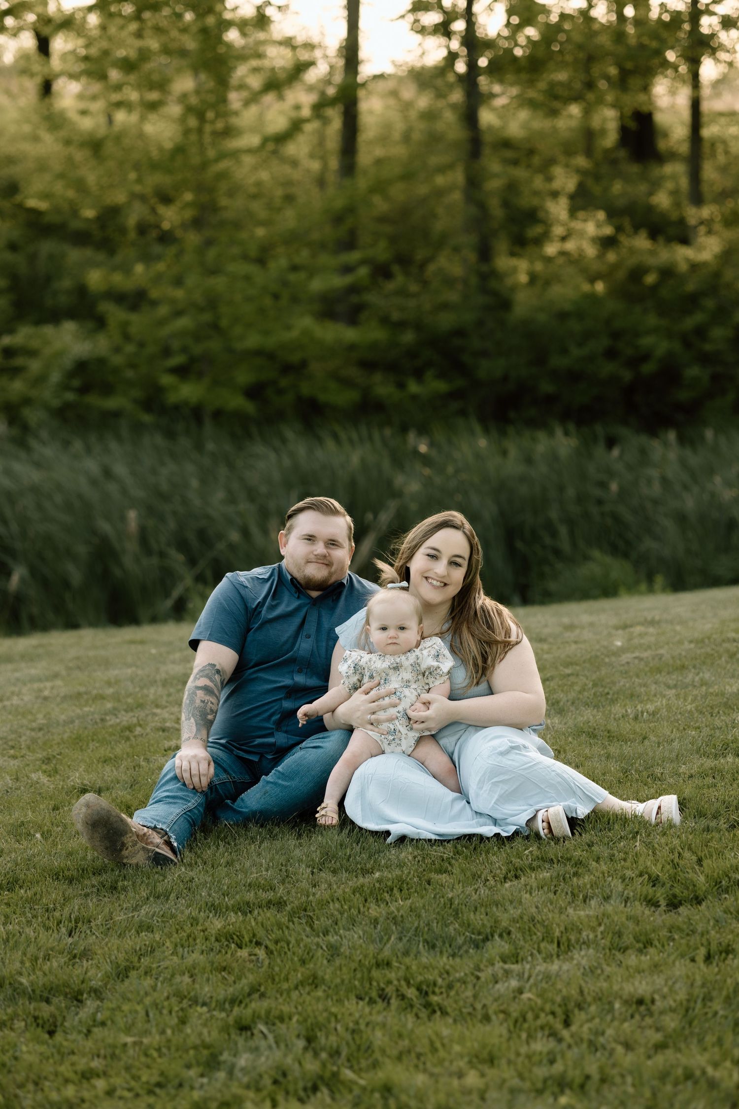 A happy family of three sits together on green grass, smiling at the camera during an outdoor photo session.