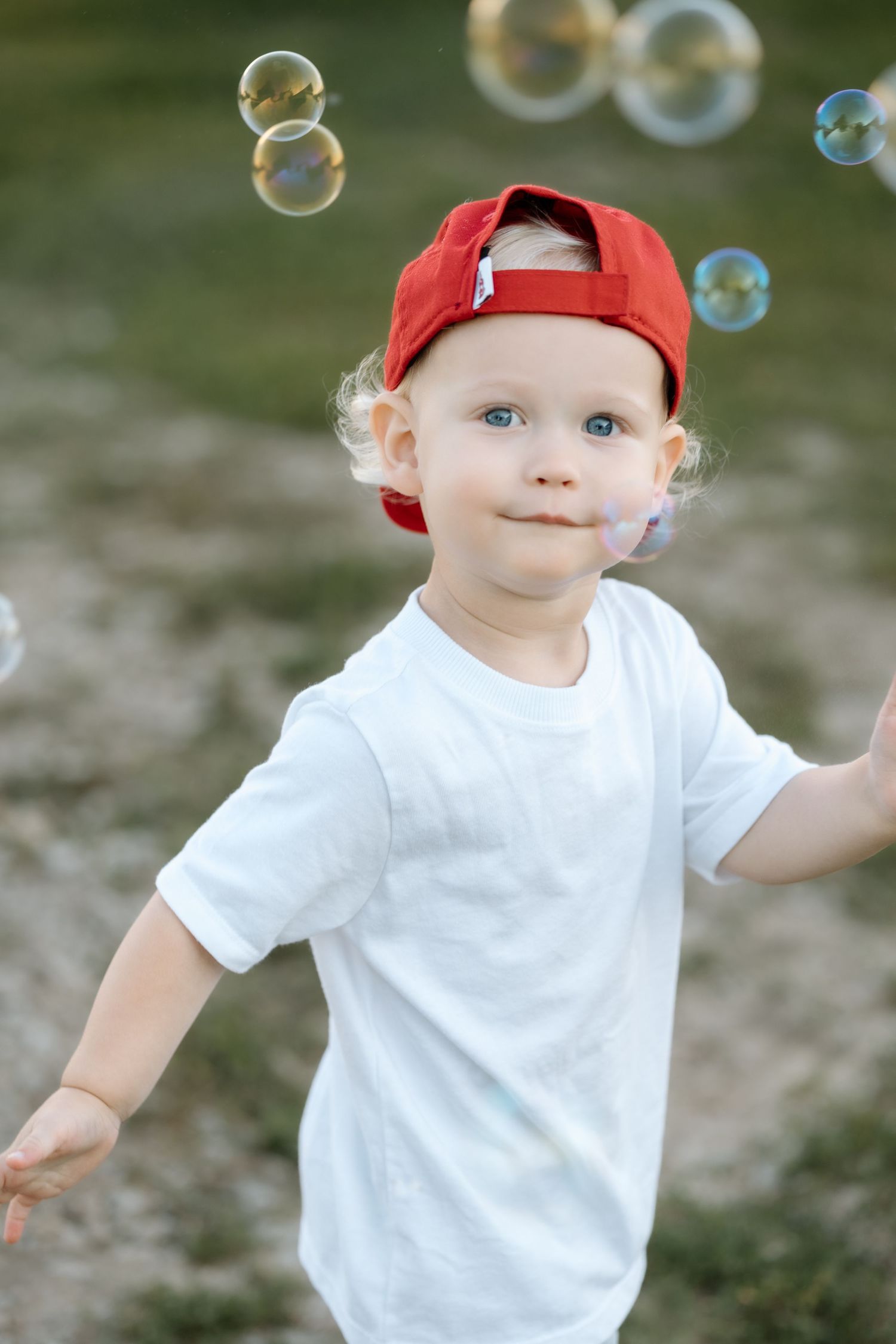 Adorable blond toddler boy in red cap and white shirt surrounded by floating soap bubbles.