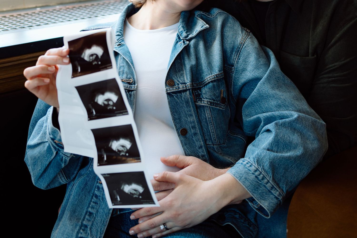 Pregnant woman in denim jacket holding ultrasound photos over baby bump, close-up view.