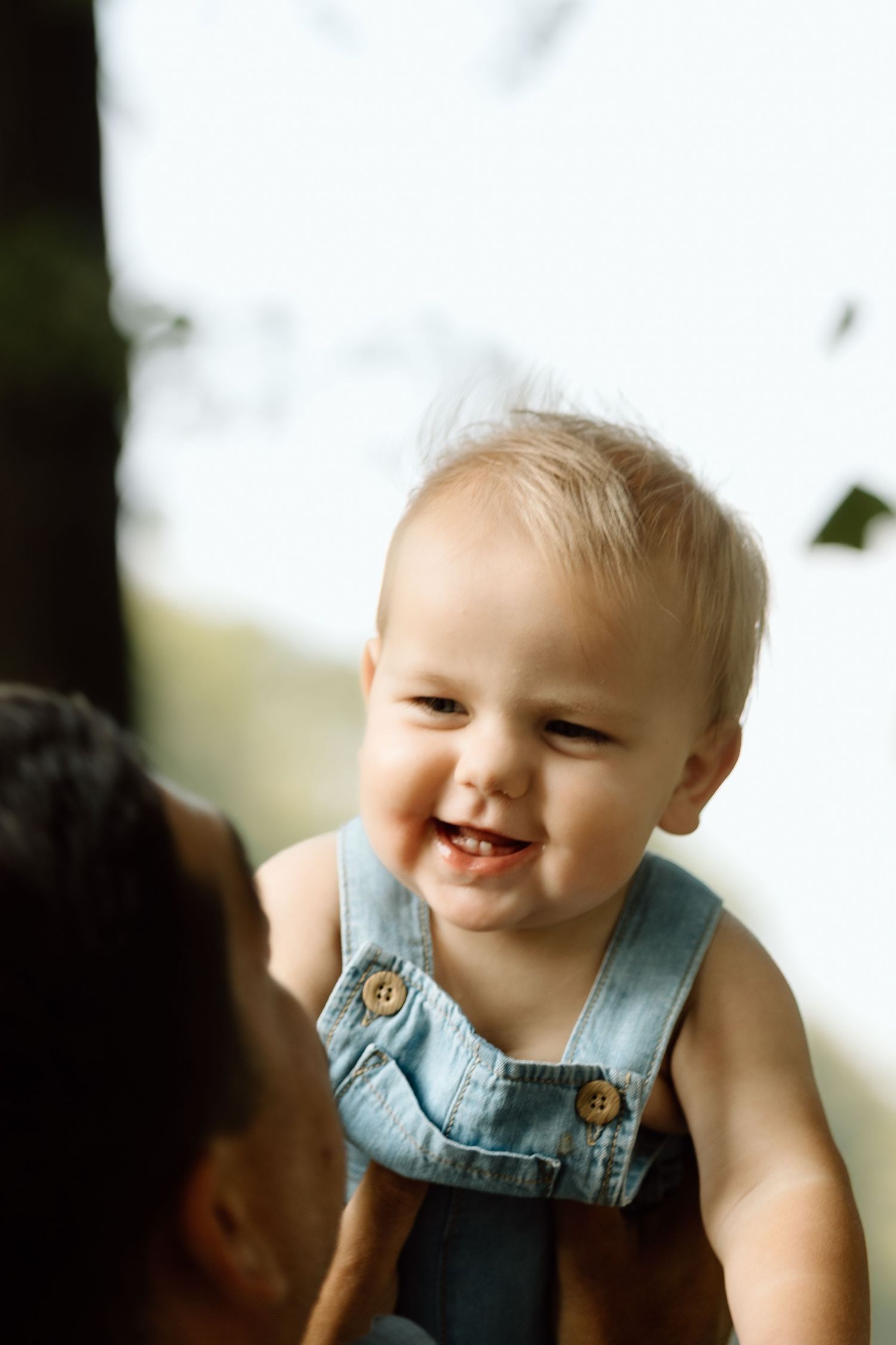 Laughing baby in denim overalls being held up by parent in sunlit outdoors.