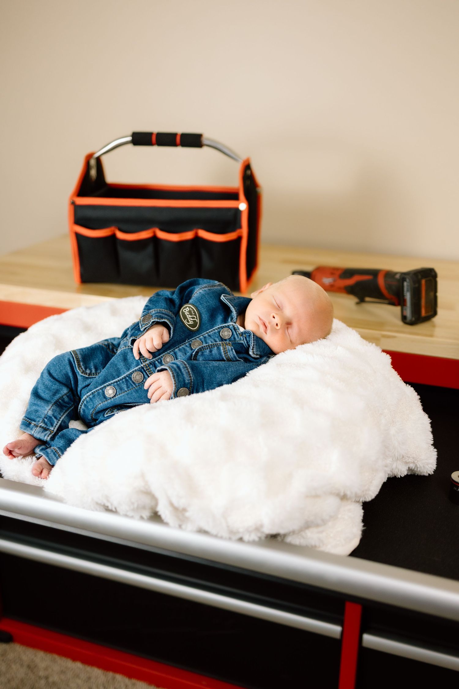 Sleeping newborn in denim outfit lying on white fur blanket beside a tool bag and drill.