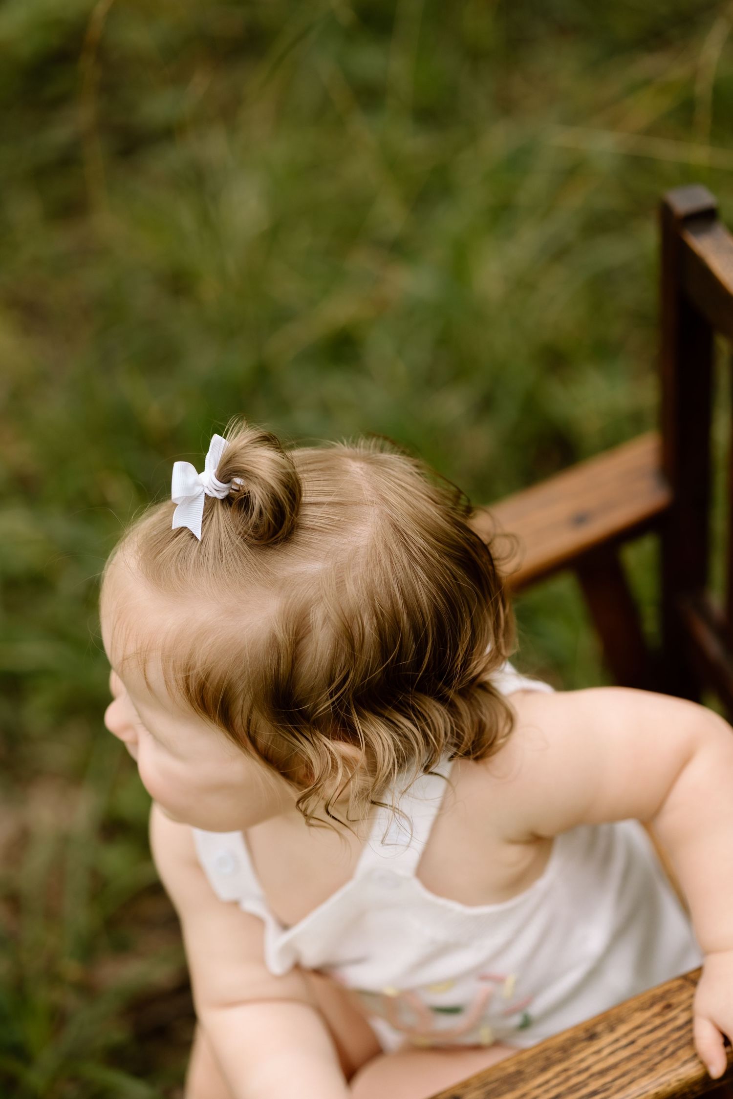 Overhead view of baby girl with white bow hair clip sitting on wooden chair in grass.