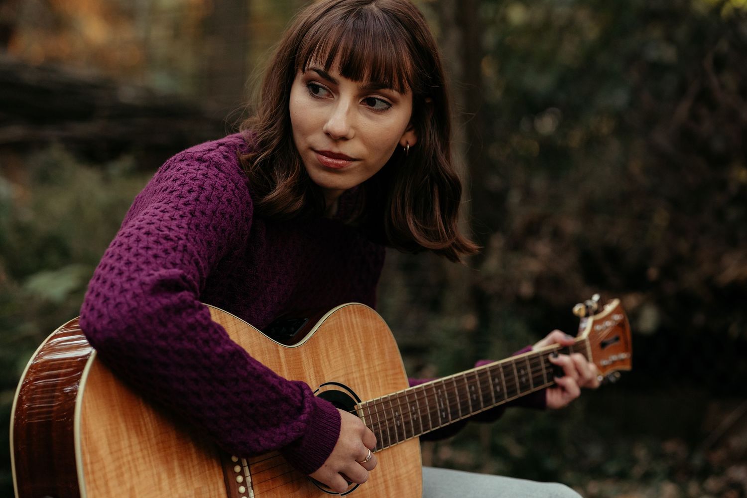 Brunette woman in purple knit sweater holds acoustic guitar while looking at camera outdoors.