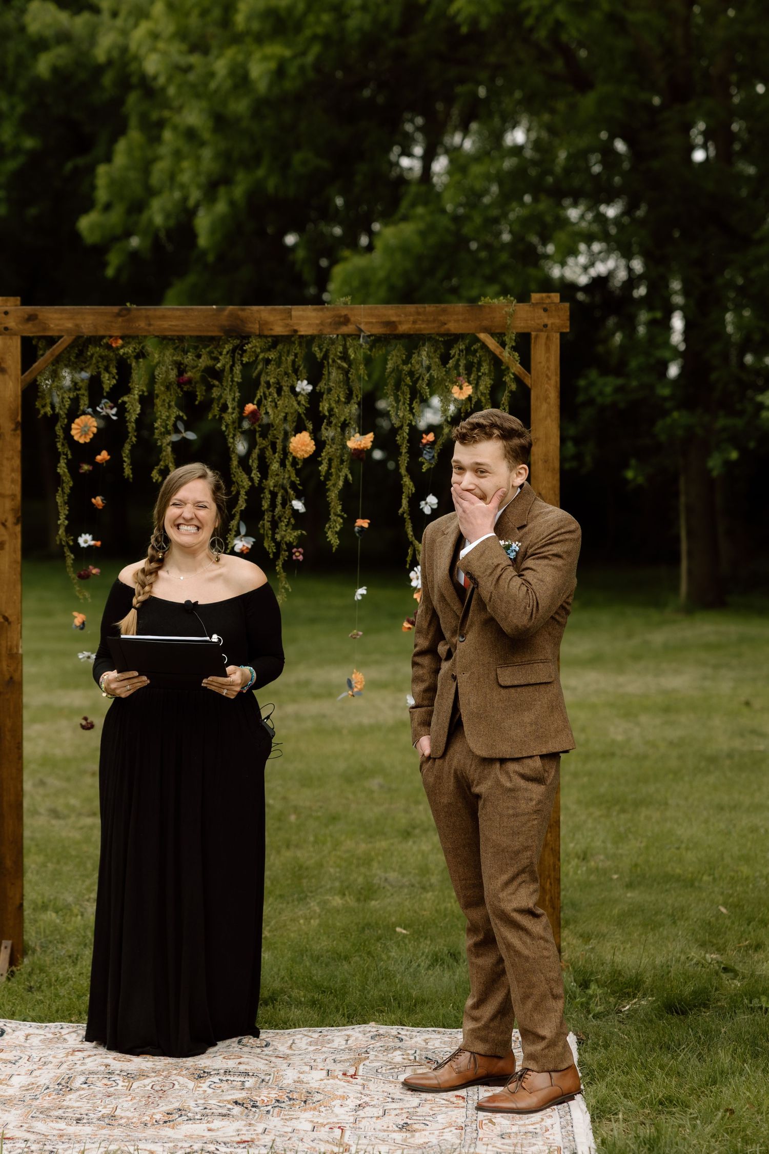 Groom laughs joyfully during outdoor wedding ceremony beside officiant at wooden floral arch on green lawn.