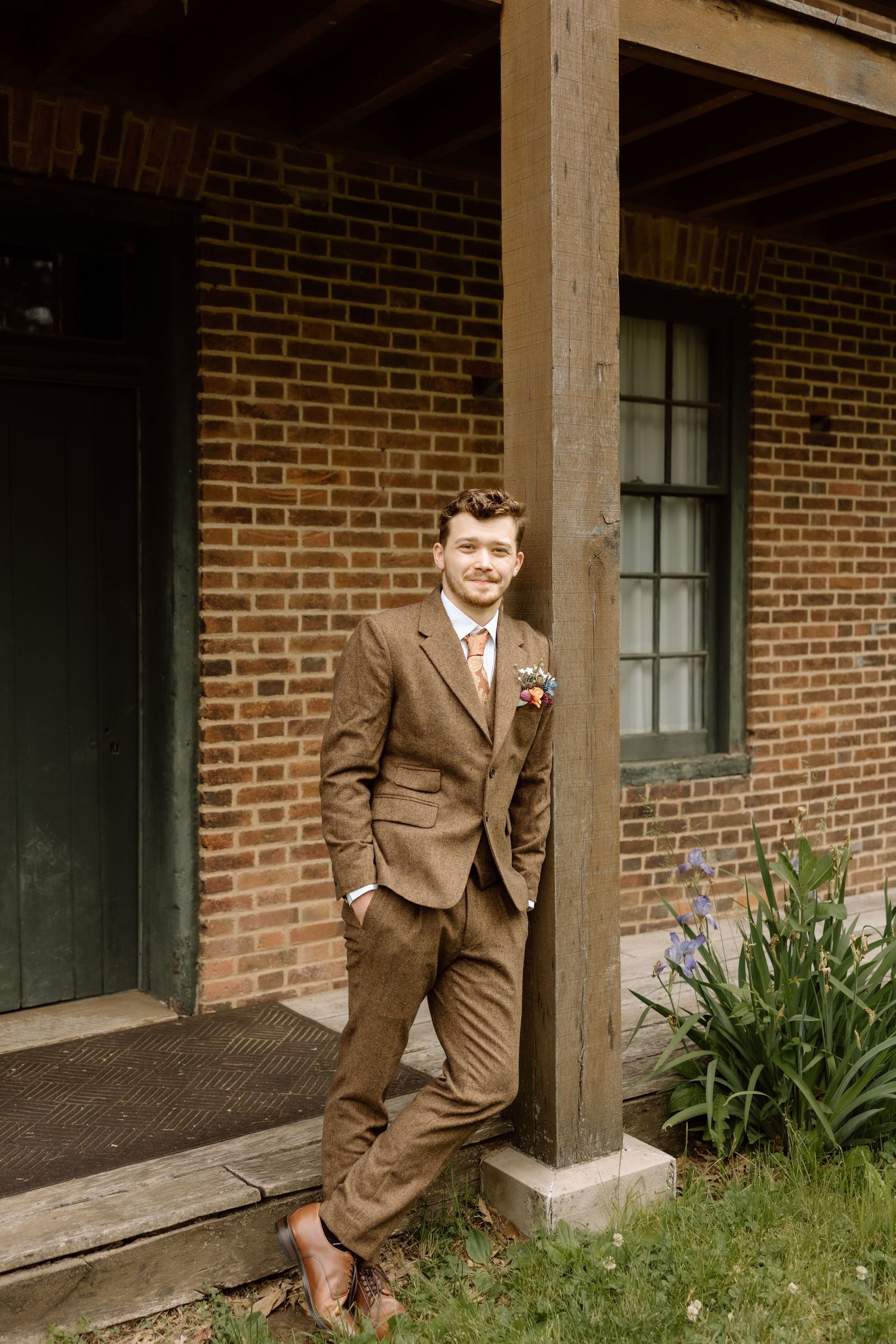 Groom in brown tweed three-piece suit with floral boutonniere leaning against wooden pillar of brick building.