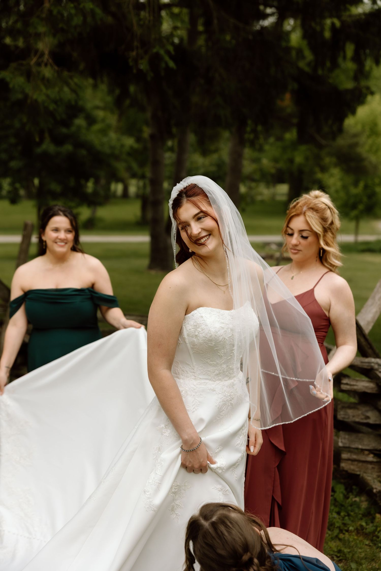 Laughing bride in lace wedding gown and veil surrounded by bridesmaids in colorful dresses outdoors.