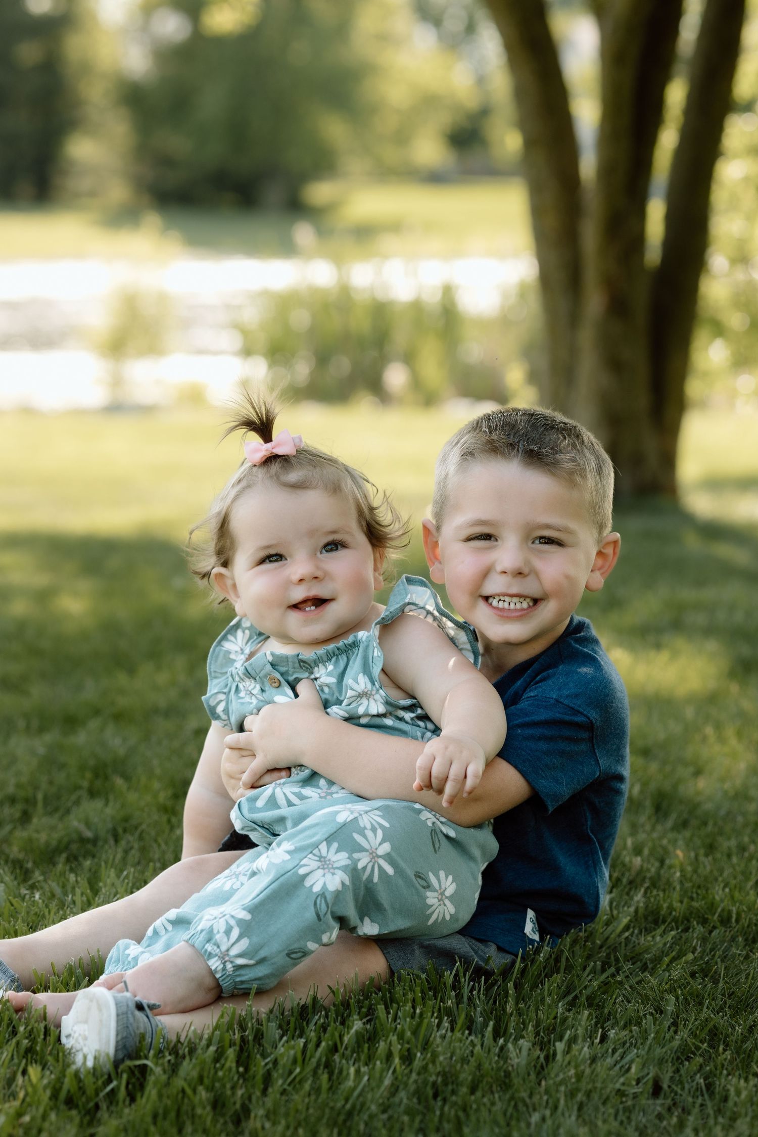 Smiling young boy hugs his baby sister in a daisy romper while sitting on green grass for sibling portraits.