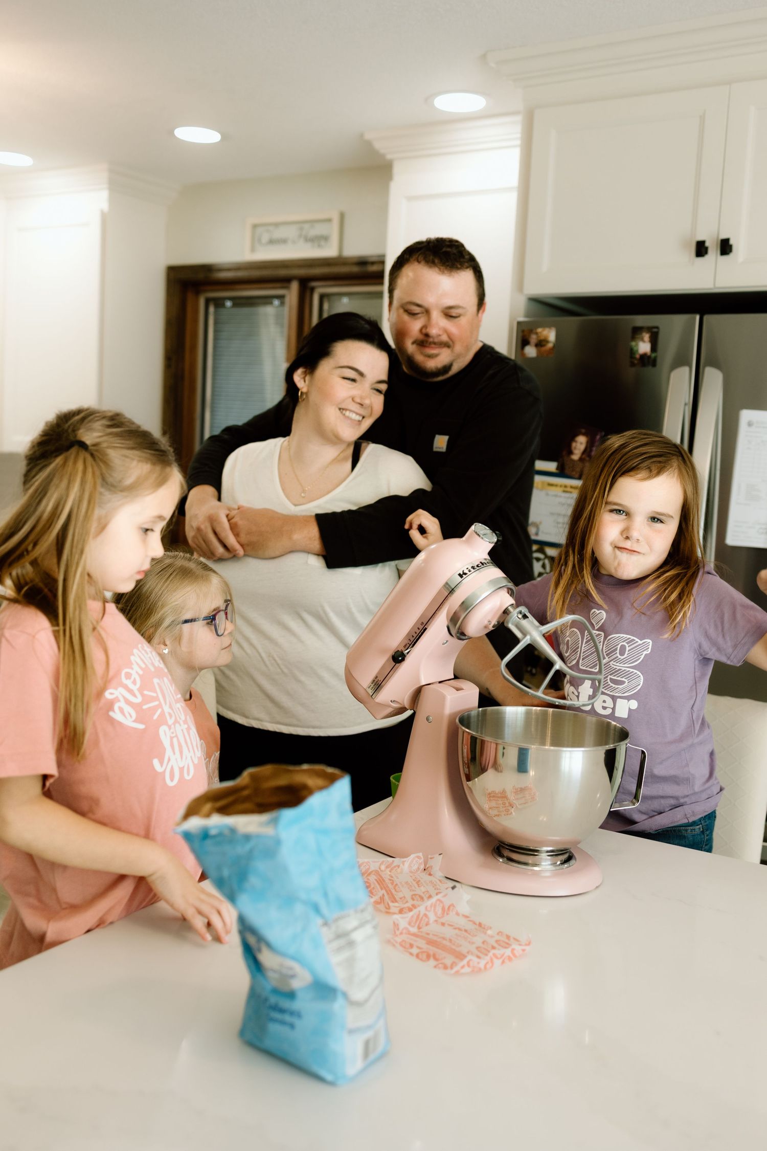 Family of five gathered around a kitchen counter using a pink KitchenAid mixer to bake together.