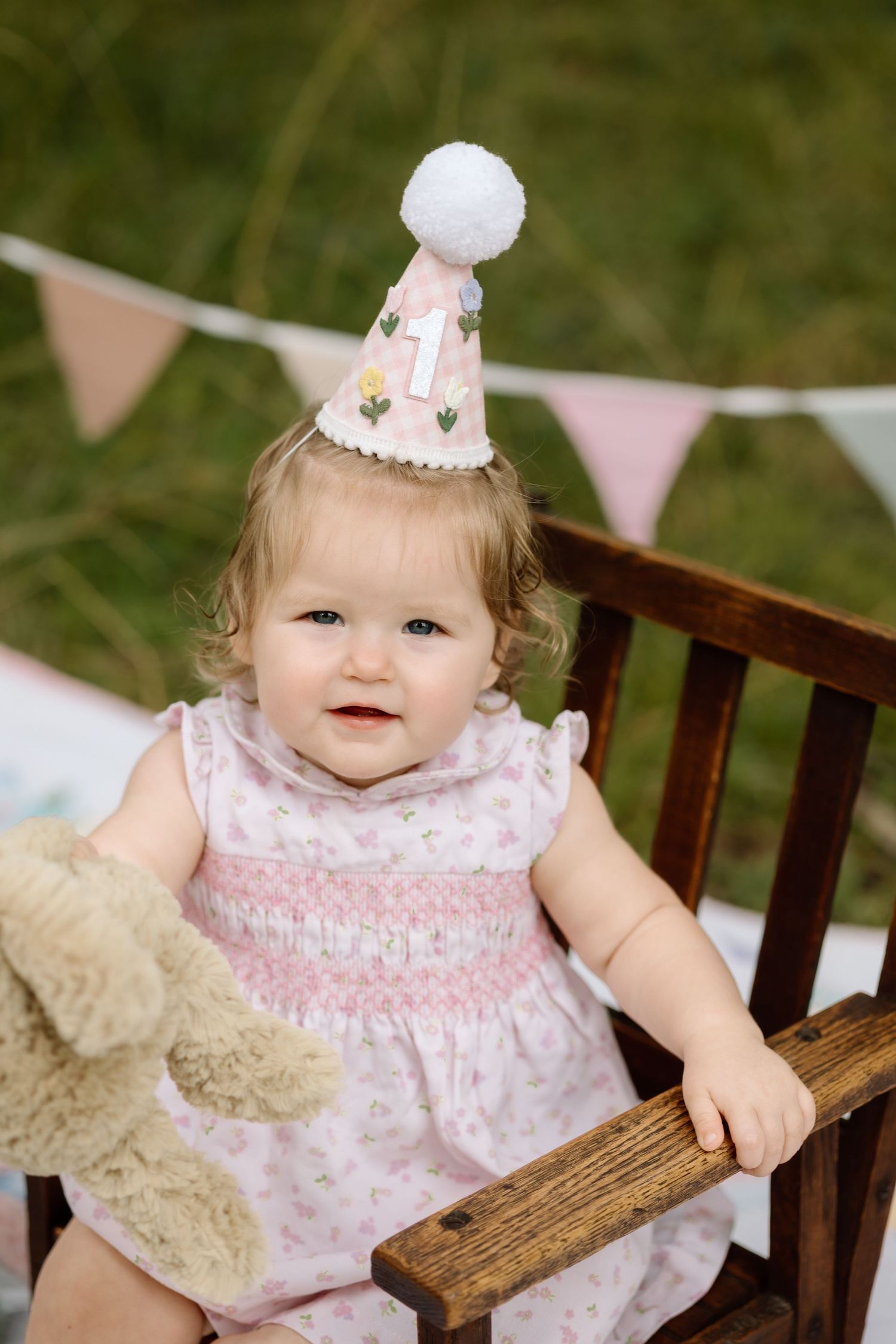 Smiling baby girl in pink smocked dress wearing first birthday party hat outdoors.