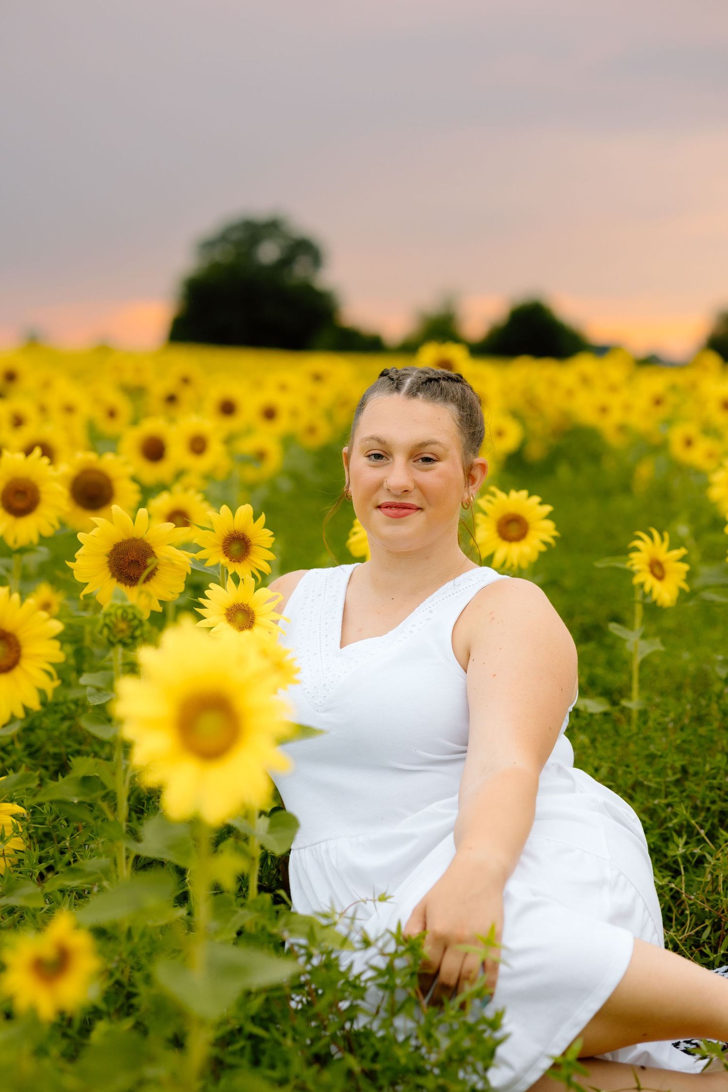 Smiling woman in white dress sitting among blooming sunflowers in a field at sunset.