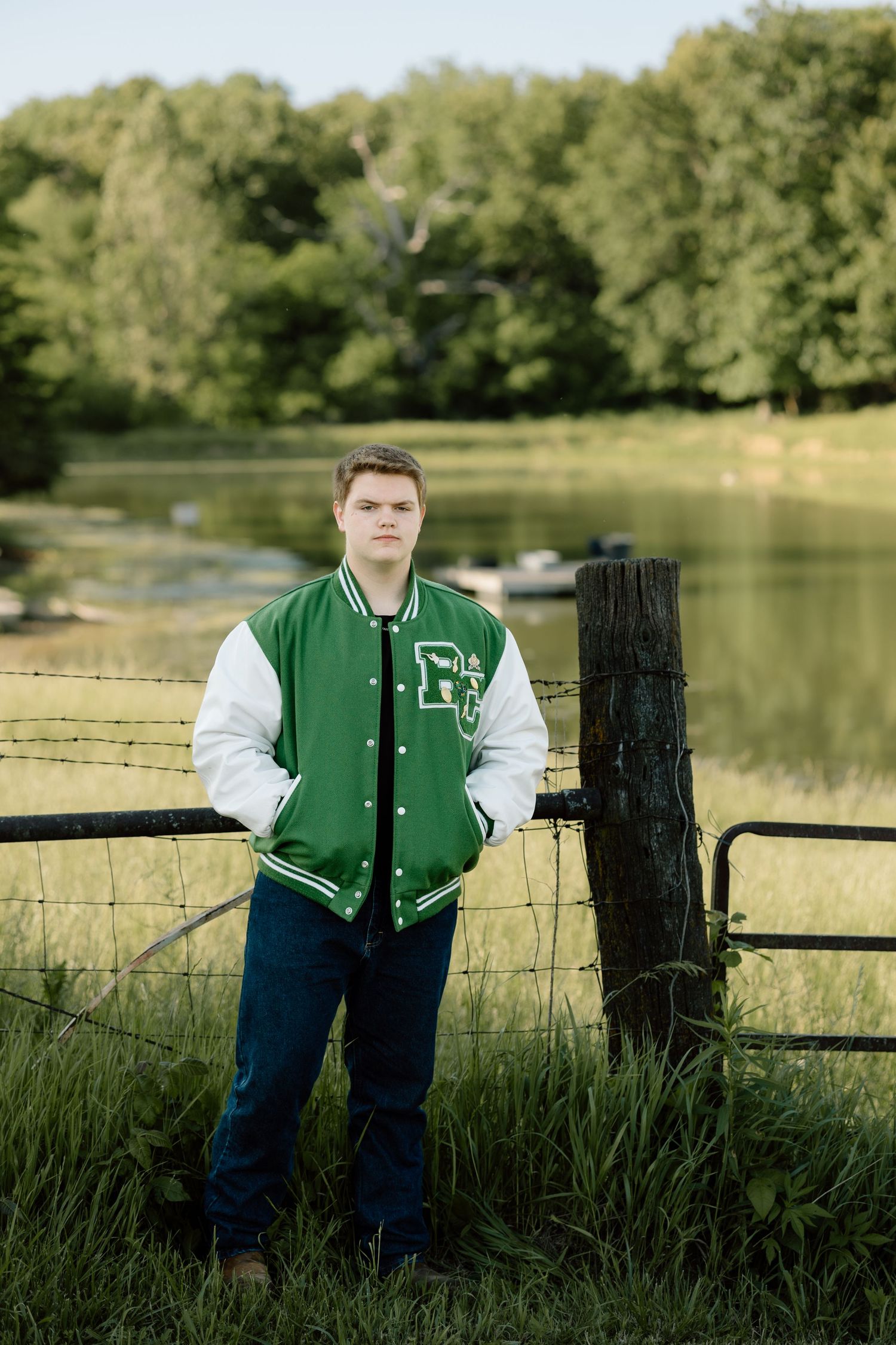 Young man in green varsity jacket leaning on a fence post near a peaceful pond.