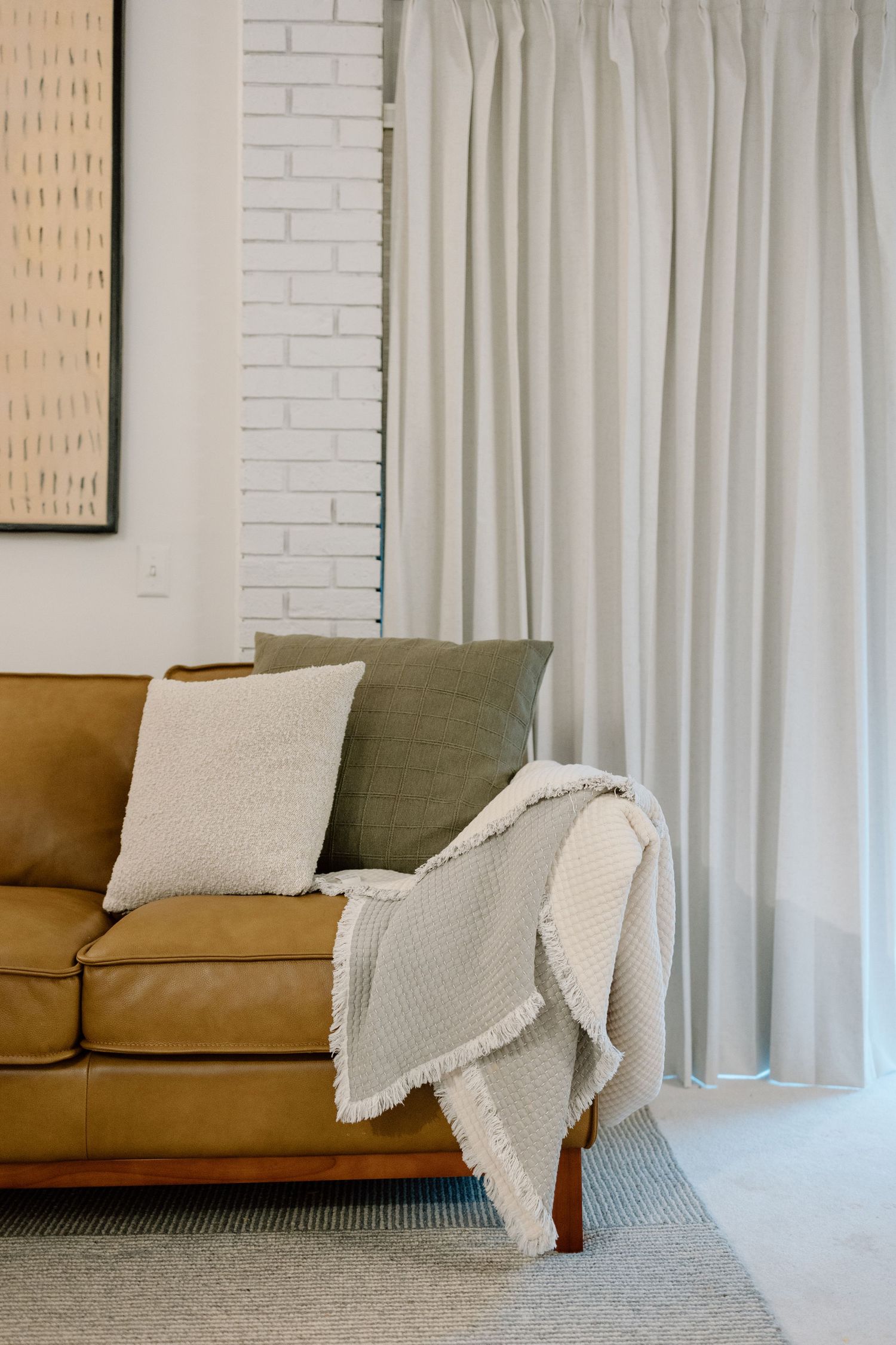 Tan leather sofa with neutral pillows and a white fringed throw blanket beside floor-length curtains.