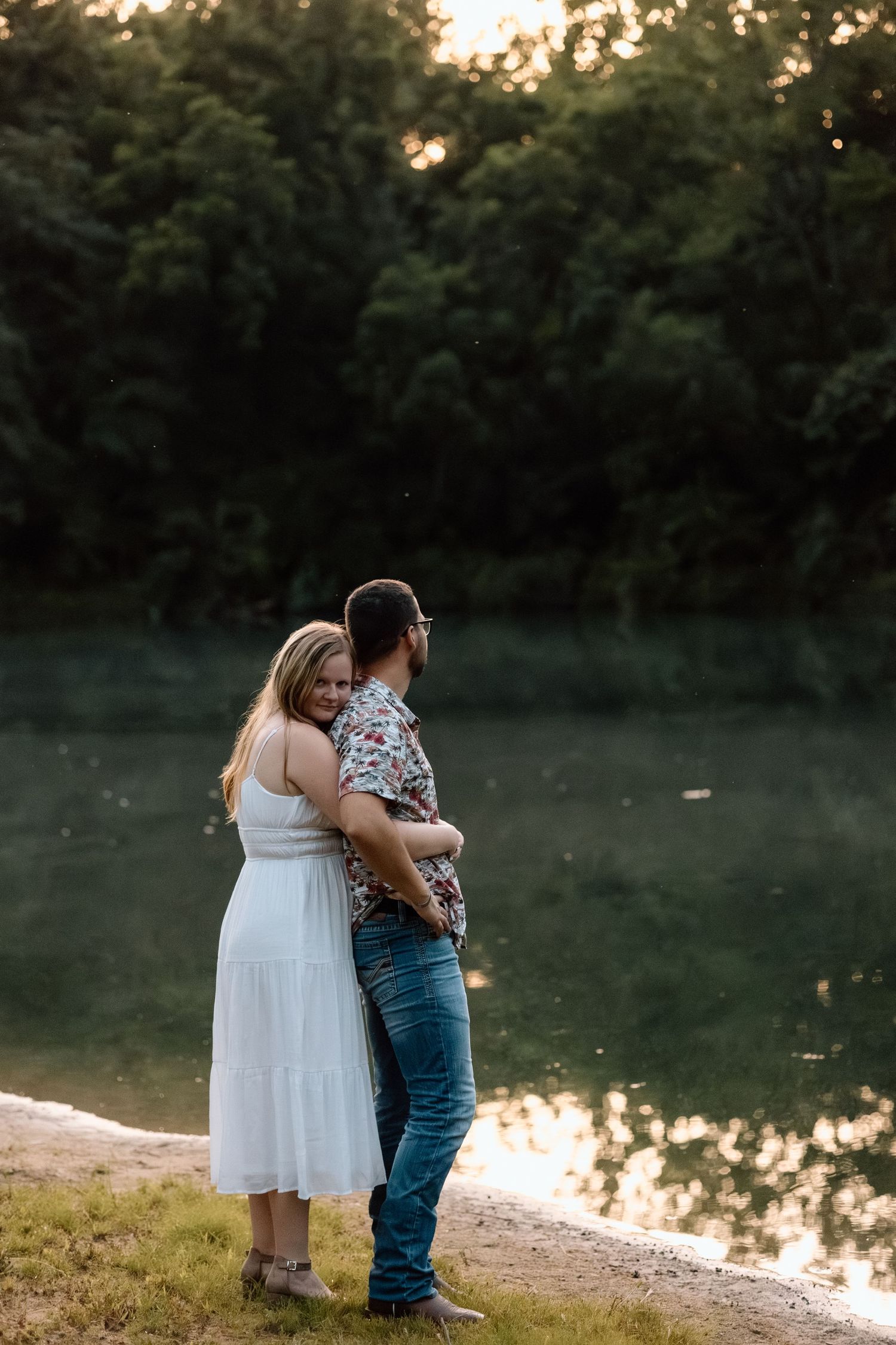 A couple embraces by a peaceful lake at golden hour, the woman in a white dress hugging the man from behind.