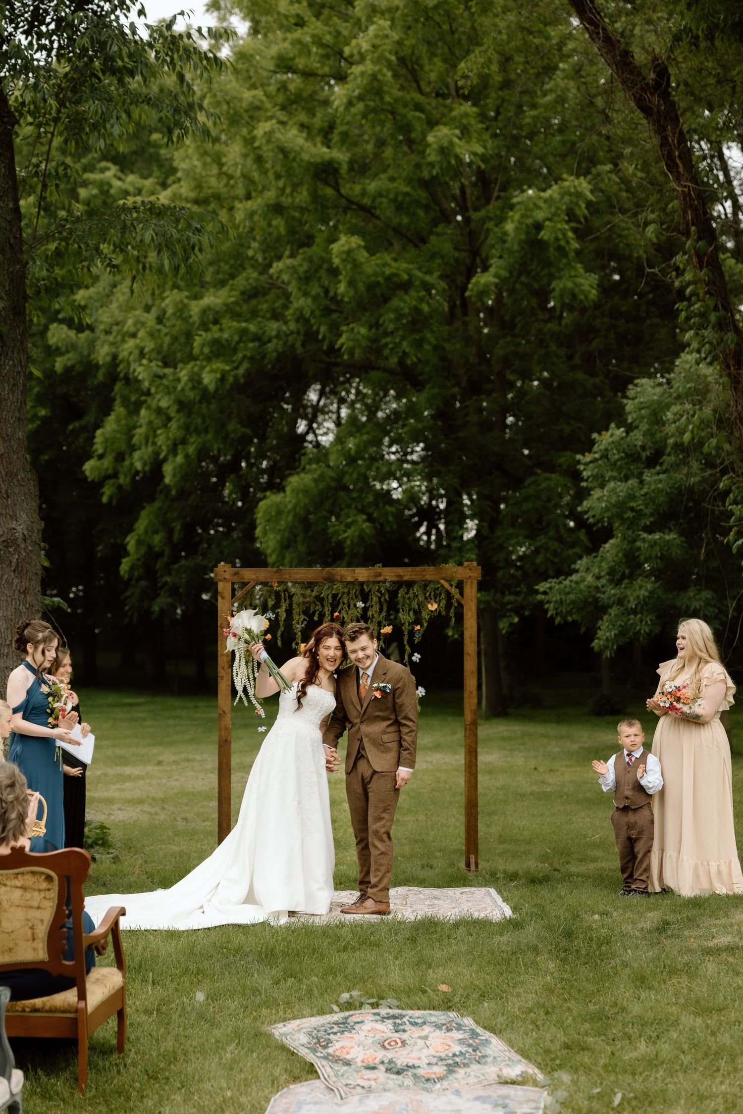 Newlyweds walk joyfully down the aisle after outdoor ceremony beneath wooden arch surrounded by wedding party.