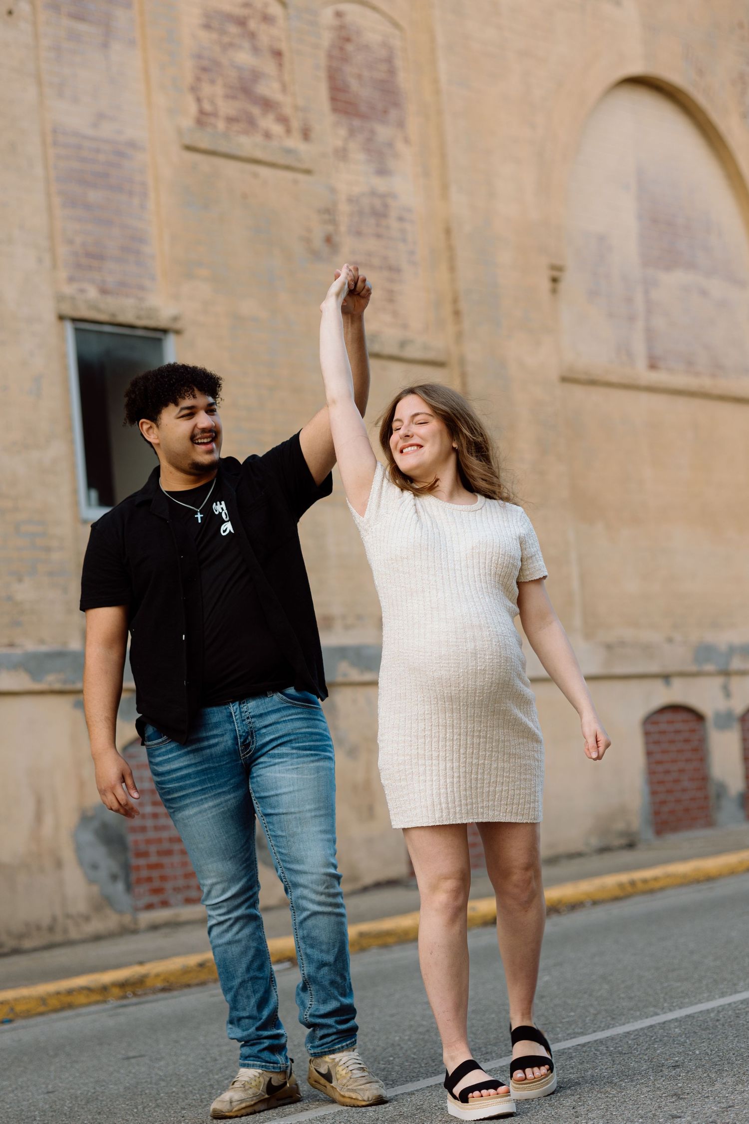 Happy pregnant couple holding hands and dancing outdoors near a historic brick building on a sunny day.