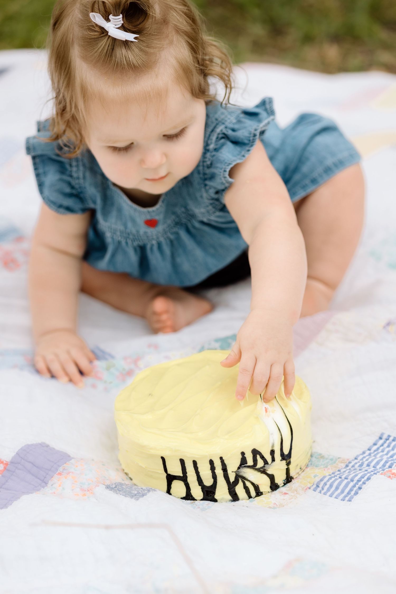 Baby girl in denim dress reaching to touch yellow first birthday smash cake on quilt outdoors.