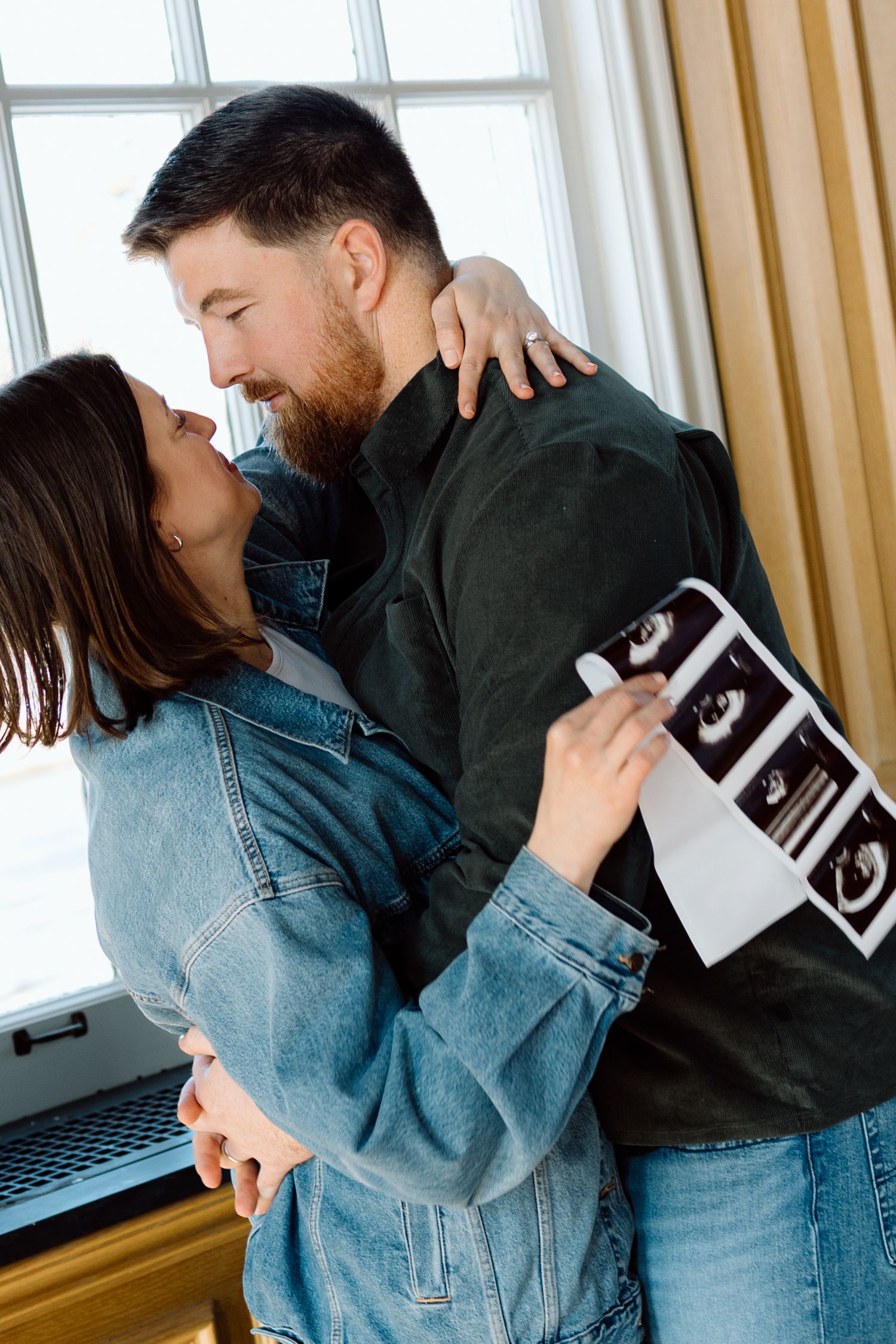 Expecting couple embraces near window while holding ultrasound photos during pregnancy announcement session.