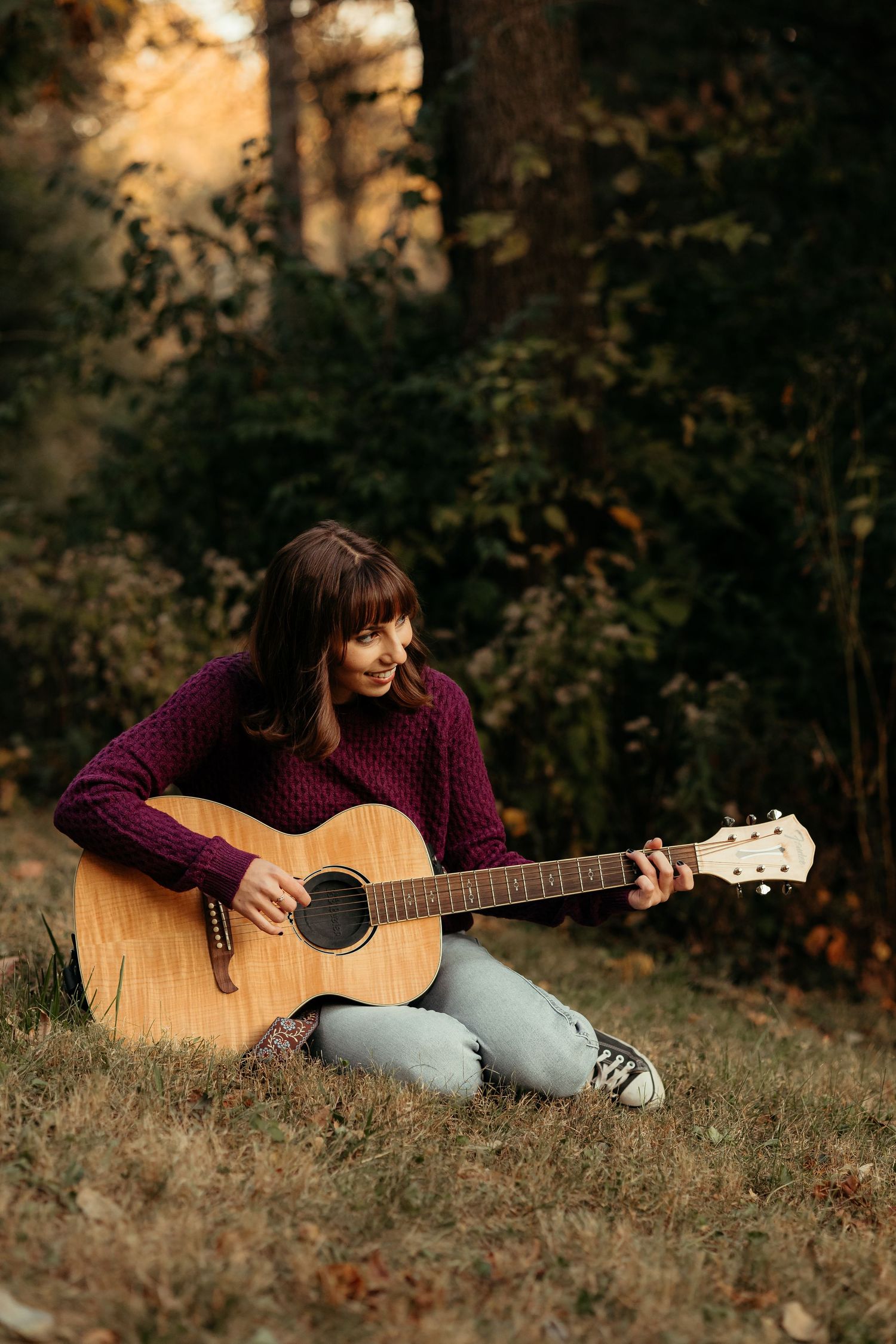 Smiling woman in purple sweater plays acoustic guitar while sitting on grass in autumn forest.