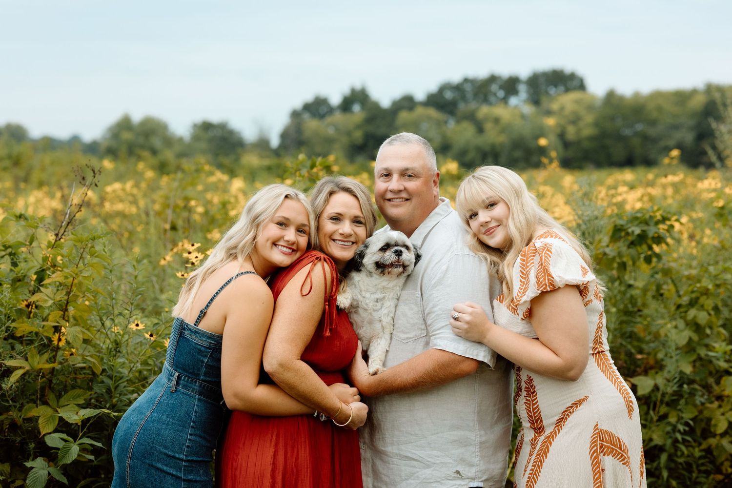 Family of four smiling together in a sunny wildflower field while holding a small Shih Tzu dog.