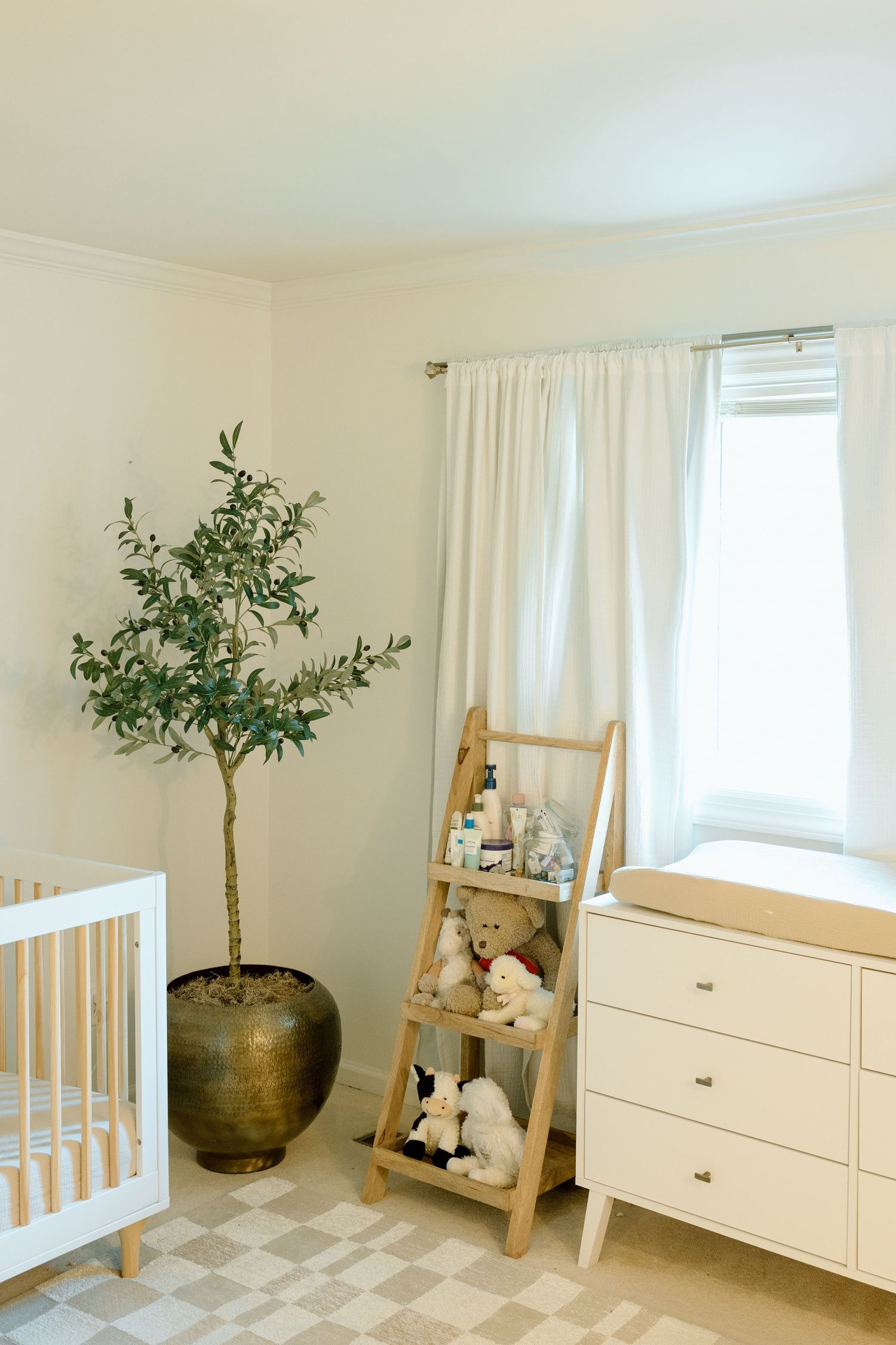 Neutral nursery with white crib, olive tree, ladder shelf, and checkered rug.