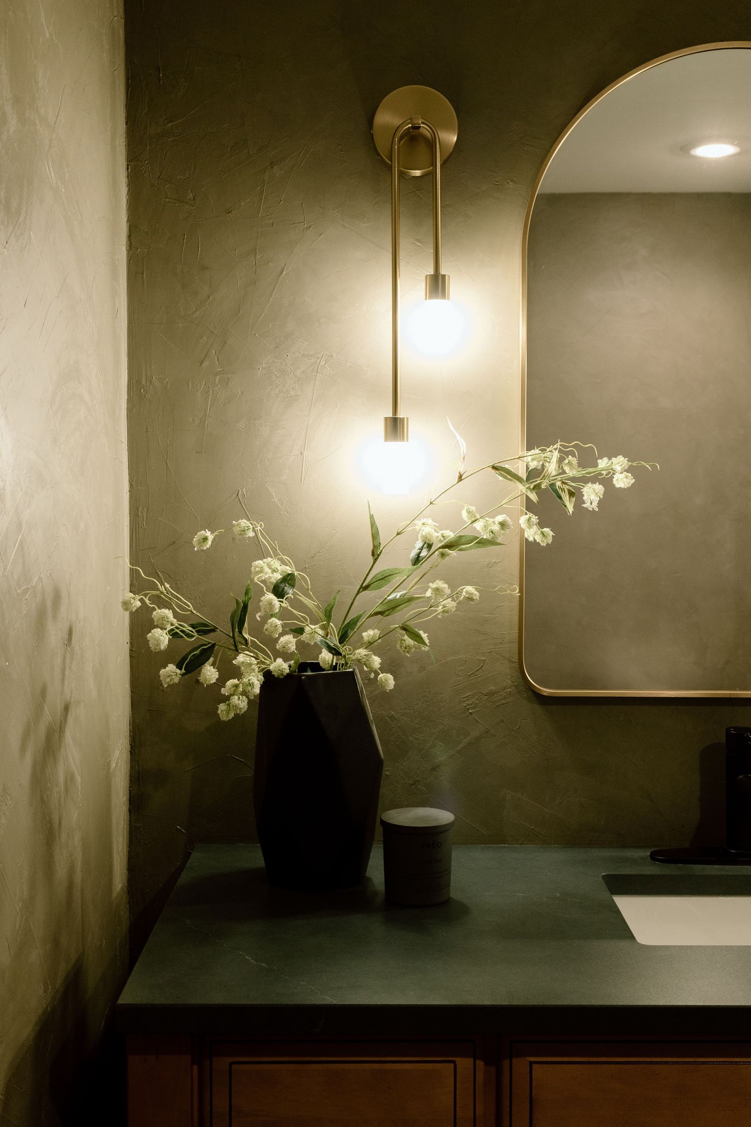 Dark vase with lily of the valley stems beside a brass sconce and arched mirror on green plaster wall.