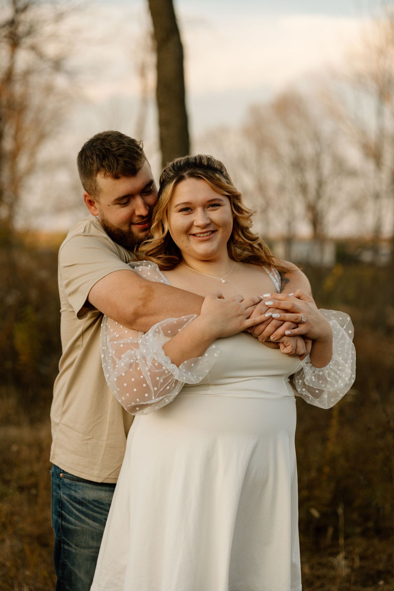 Man tenderly embraces woman in white lace dress from behind, both smiling in a sunlit autumn outdoor setting.